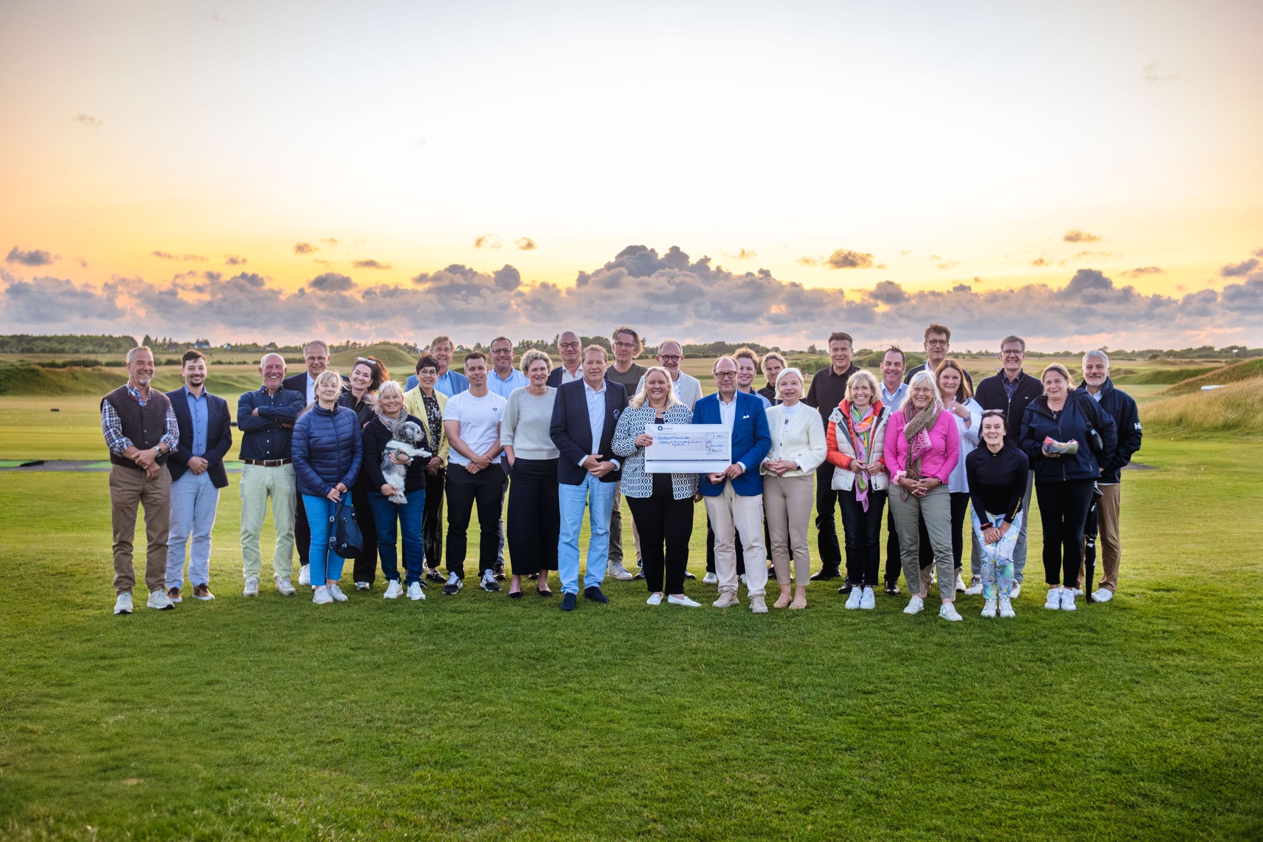 Eine große Gruppe von Menschen steht bei Sonnenuntergang auf einem grünen Golfplatz zusammen und lächelt in die Kamera; eine Person im Vordergrund hält einen großen Scheck. Der Himmel ist bunt bewölkt.