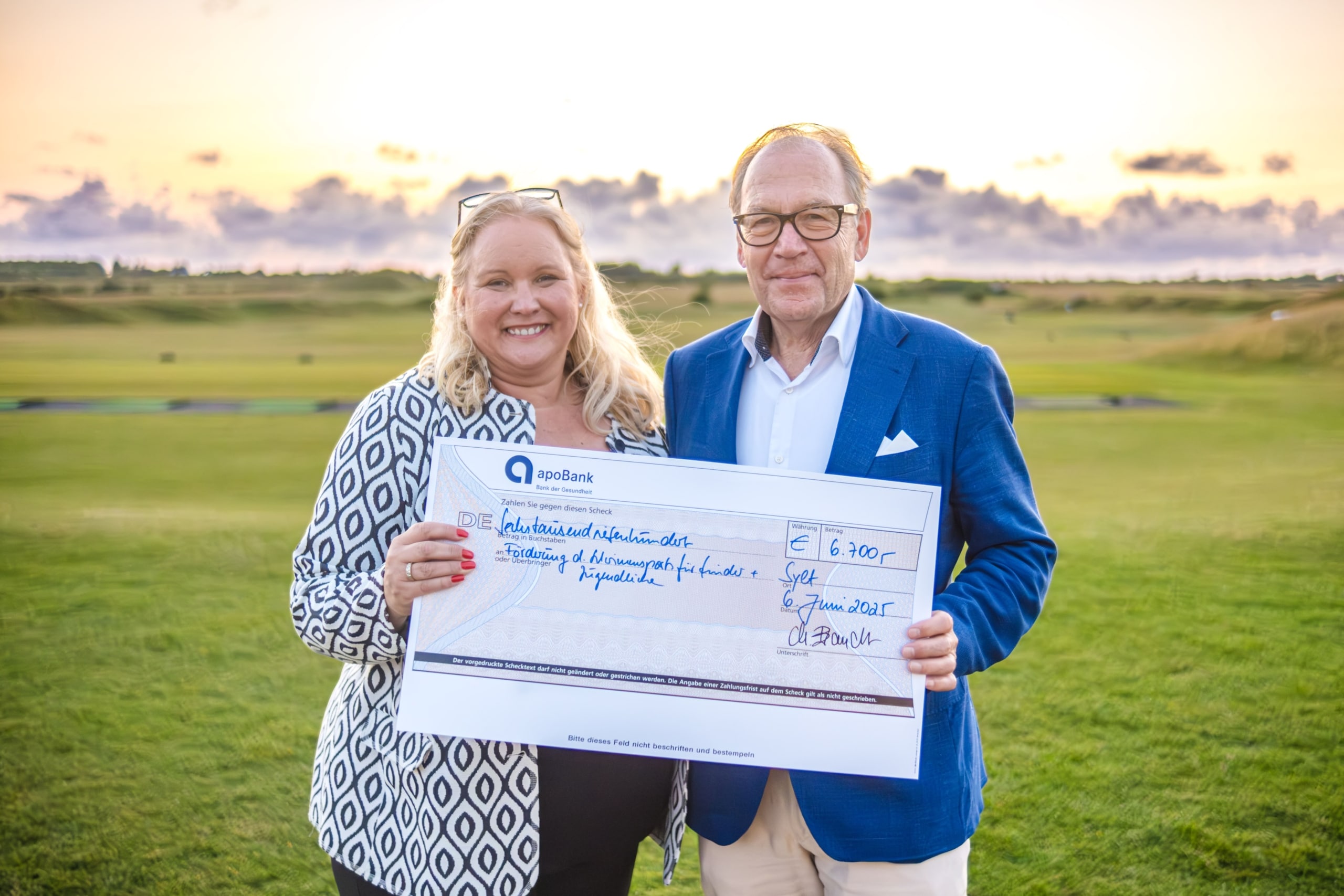 Eine lächelnde Frau und ein lächelnder Mann stehen auf einem Golfplatz und halten einen großen feierlichen Scheck über 6.900 € von den Sparekassen Vendsyssel in der Hand, mit einem malerischen Sonnenuntergang im Hintergrund.