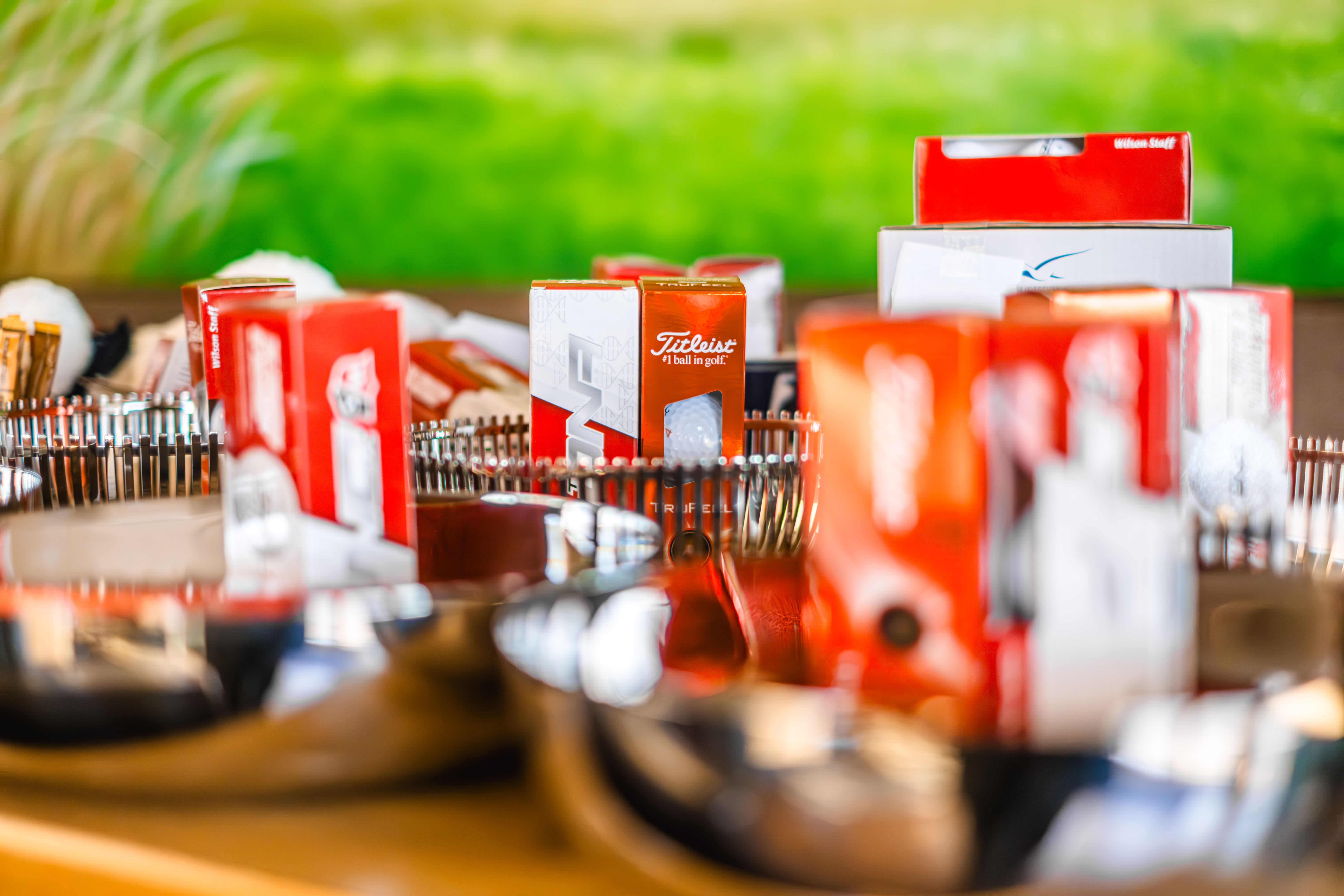 In mehreren silberglänzenden Tabletts stapeln sich rote und weiße Titleist-Golfballschachteln vor einem verschwommenen grünen und gelben Hintergrund.