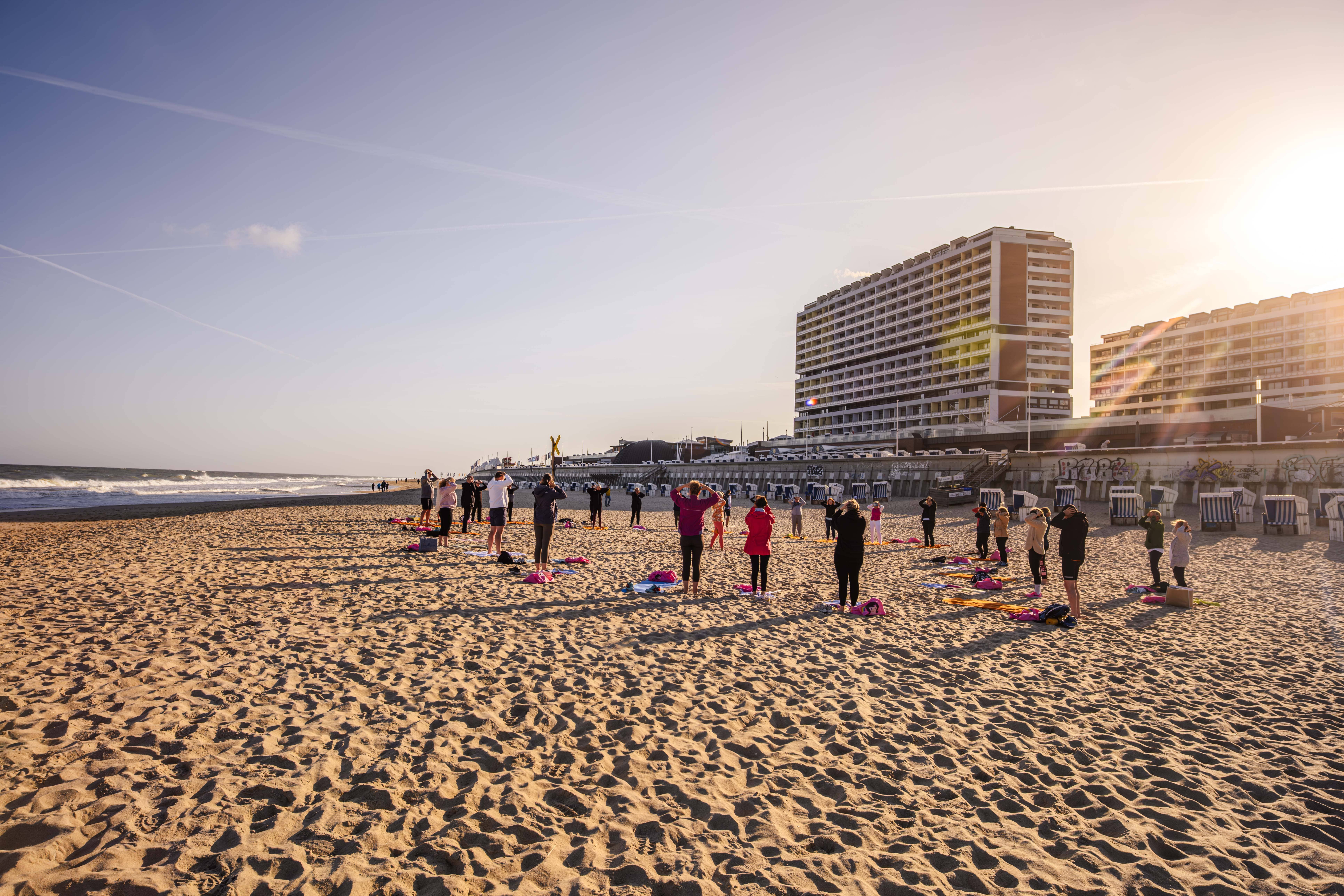 Eine Gruppe von Menschen praktiziert Yoga an einem Sandstrand bei Sonnenuntergang in der Nähe des Ozeans, mit hohen modernen Gebäuden im Hintergrund und Sonne, die von der rechten Seite des Bildes scheint.