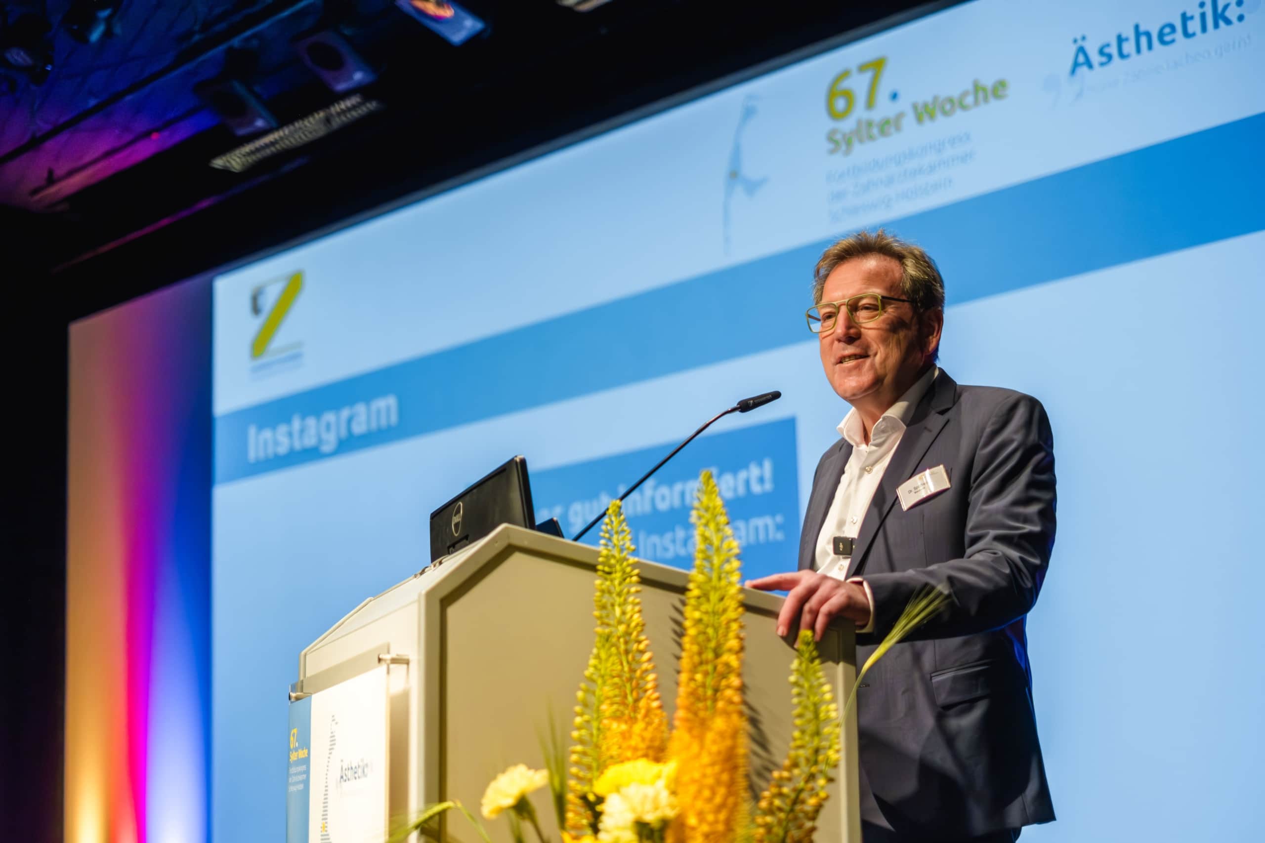 Ein Mann im Anzug spricht während der 67. Sylter Woche auf einem Podium auf der Bühne. Auf einem großen Bildschirm hinter ihm werden Veranstaltungsdetails und das Wort Instagram angezeigt. Gelbe Blumen schmücken das Podium.
