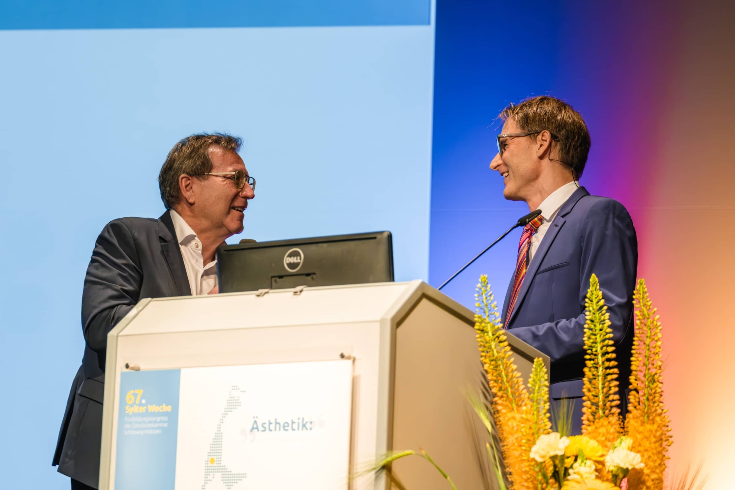 Zwei Männer in Anzügen stehen an einem Podium und lächeln sich während einer formellen Veranstaltung zu. Auf dem Podium steht ein Schild mit Text und davor sind Blumen angeordnet. Der Hintergrund weist einen blauen und orangefarbenen Farbverlauf auf.