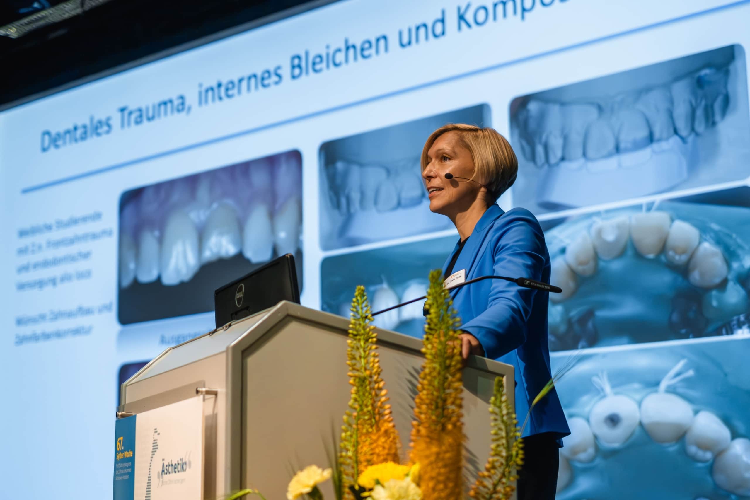 Eine Frau in einem blauen Blazer steht an einem Podium und hält einen Vortrag über Zahntraumata, während hinter ihr auf einem großen Bildschirm Bilder von Zähnen und ein deutscher Text angezeigt werden. Gelbe Blumen schmücken das Podium.