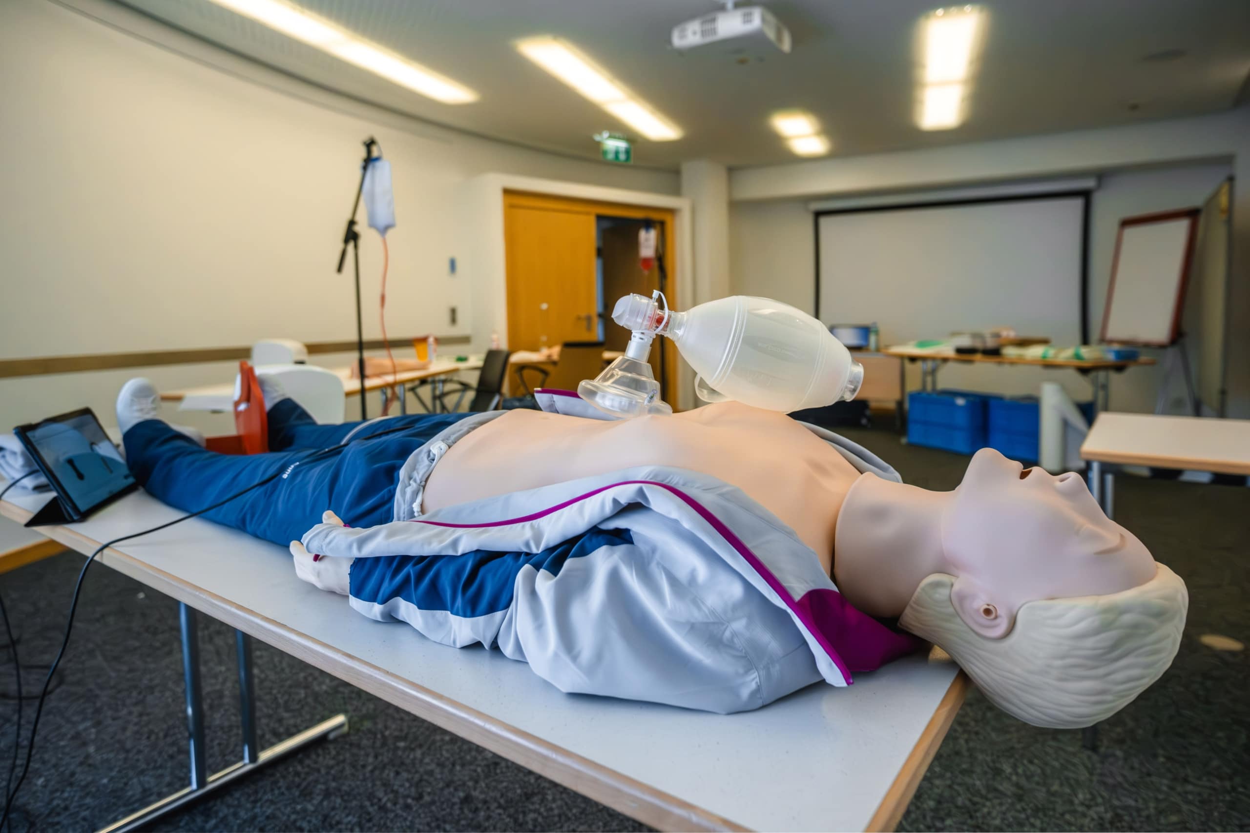 Eine Schaufensterpuppe für die Herz-Lungen-Wiederbelebung liegt auf einem Tisch in einem Klassenzimmer, auf dessen Brust ein Wiederbelebungsbeutel liegt. Der Raum ist mit Stühlen, Tischen und medizinischen Trainingsgeräten ausgestattet.