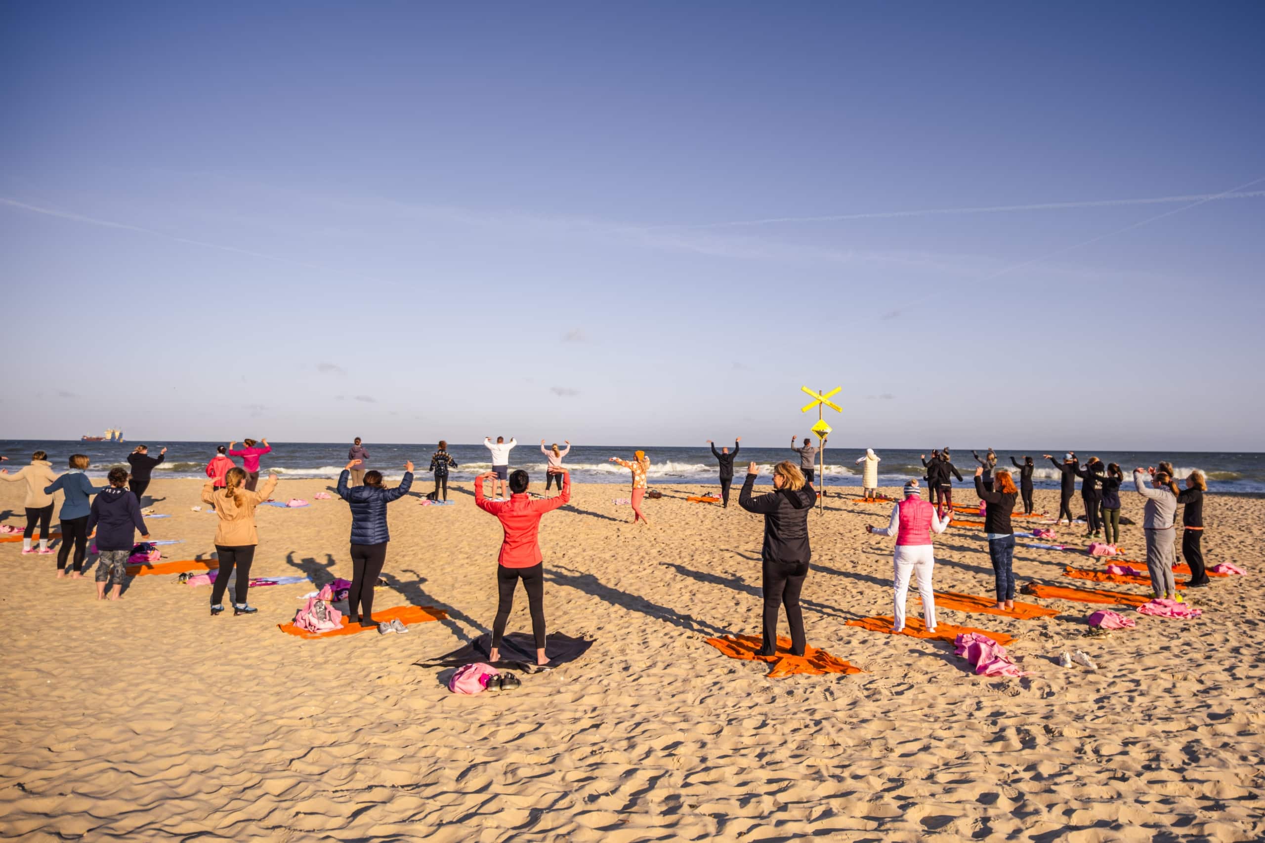 Eine Gruppe von Menschen steht an einem Sandstrand mit Blick auf das Meer und nimmt an einem Fitness- oder Yogakurs im Freien auf einzelnen Matten unter einem klaren blauen Himmel teil.