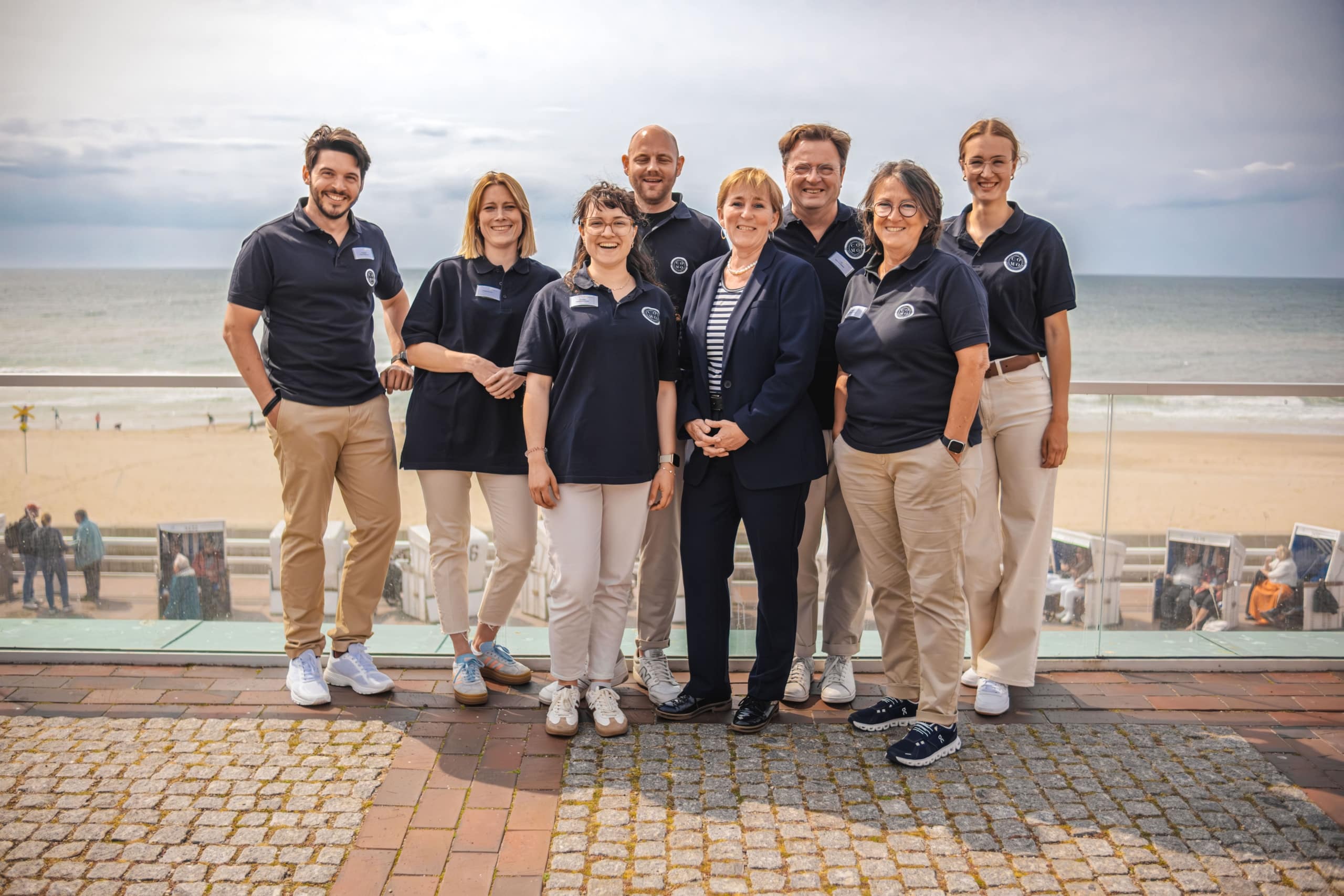Eine Gruppe von acht Personen, die meisten in marineblauen Polohemden und beigen Hosen, steht lächelnd an einer Strandpromenade mit Sandstrand und Meer im Hintergrund.