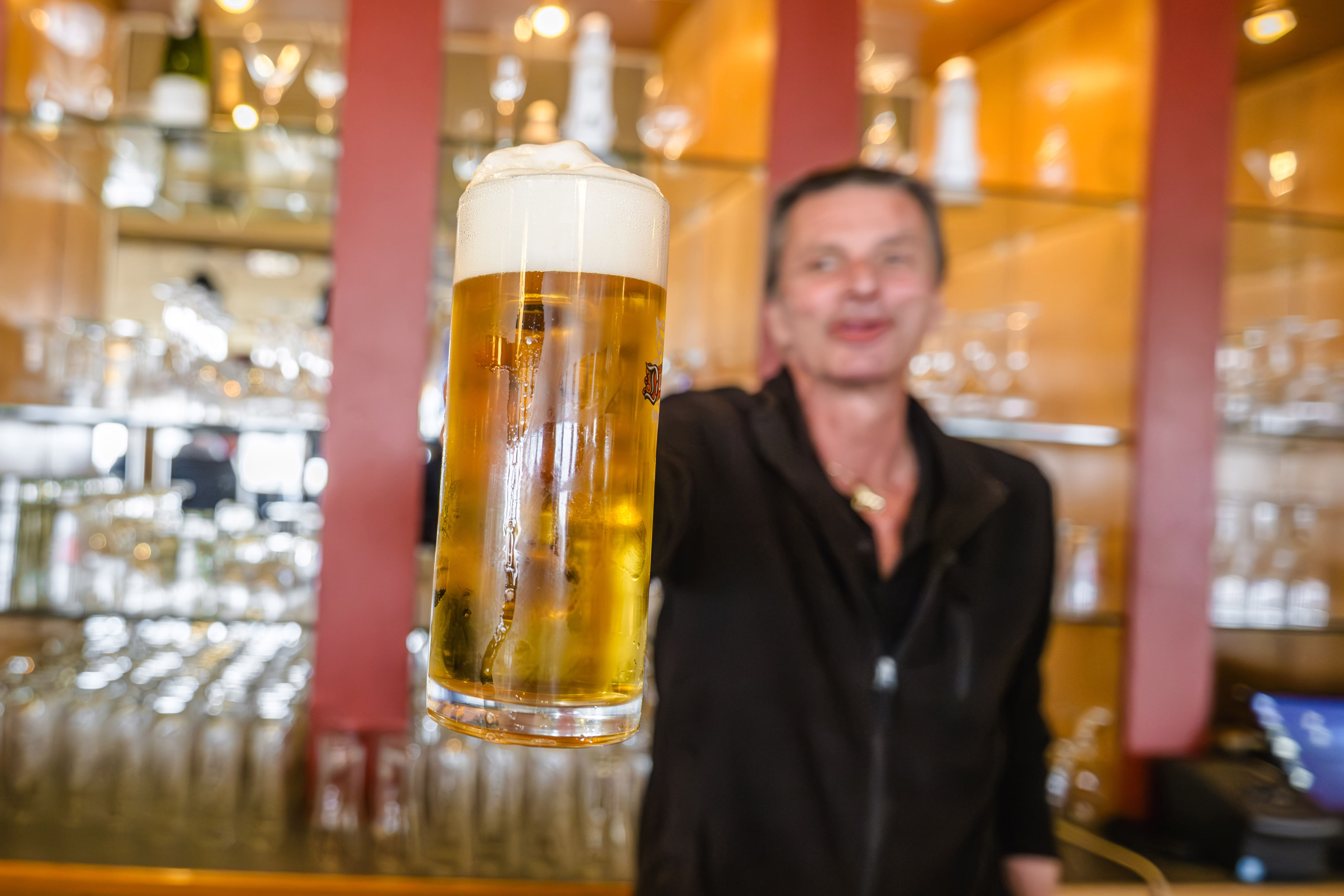 Ein Barkeeper in einem schwarzen Hemd hält ein hohes Glas Bier mit Schaumkrone in die Kamera, während er vor einem Regal mit Gläsern und Flaschen steht. Der Fokus liegt auf dem Bier, während der Barkeeper im Hintergrund verschwimmt.