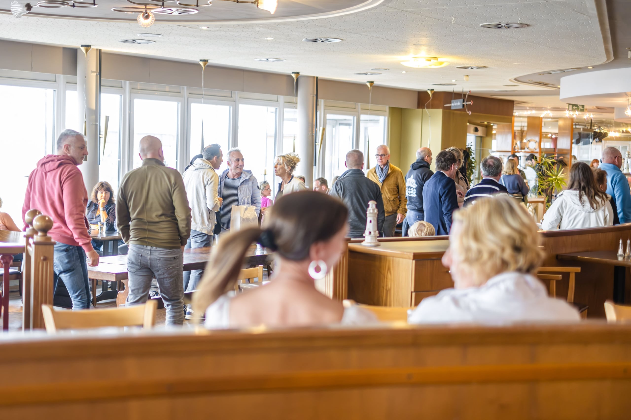 Eine Gruppe von Menschen steht und sitzt in einem hellen, modernen Restaurant mit großen Fenstern; einige unterhalten sich in Gruppen, während zwei Frauen im Vordergrund sitzen, mit dem Rücken zur Kamera.