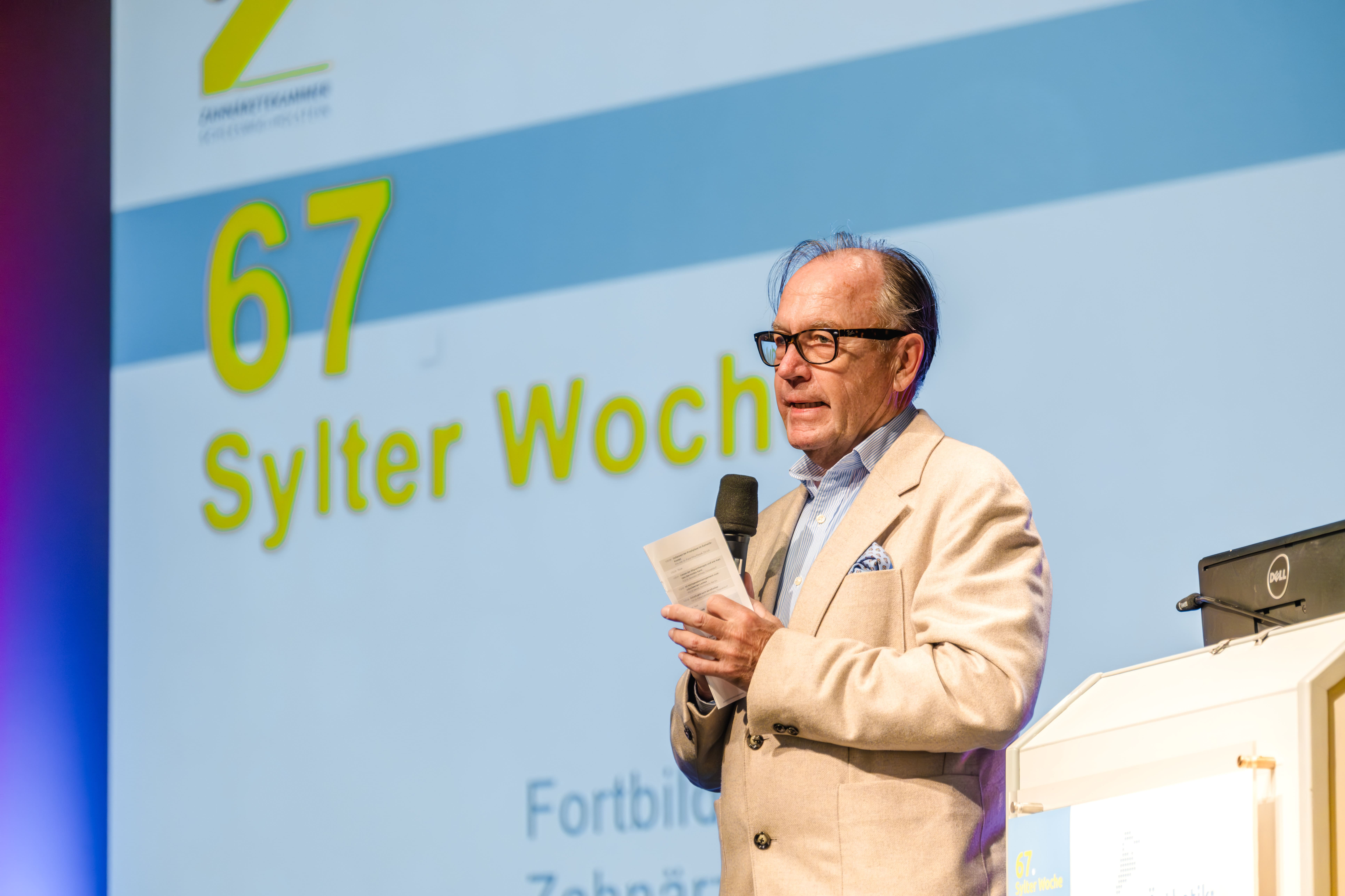 Ein Mann mit Brille und beigem Blazer spricht auf der Bühne der 67. Sylter Woche in ein Mikrofon und steht vor einem großen Bildschirm, auf dem der Name und das Logo der Veranstaltung zu sehen sind.