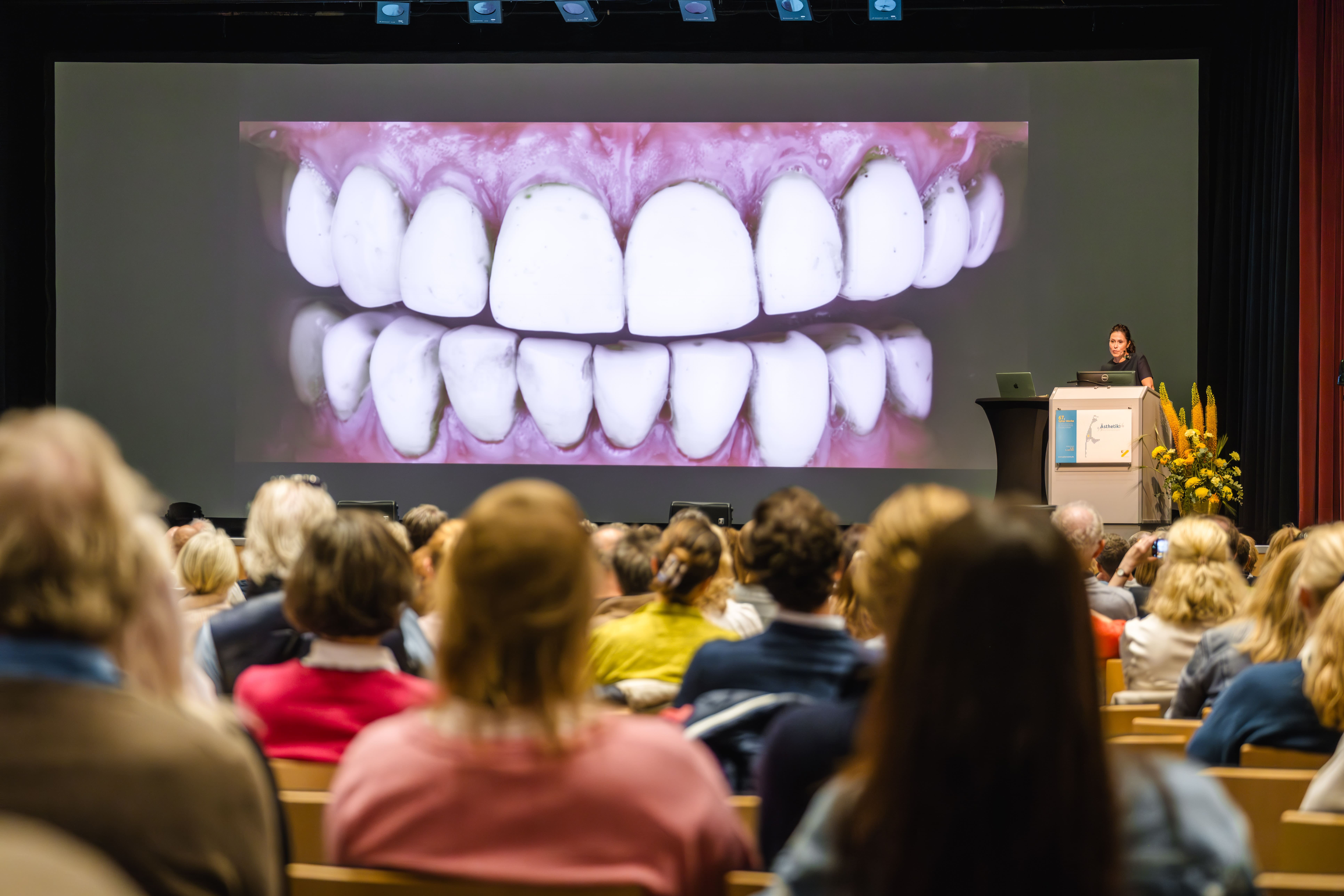 Ein großes Publikum verfolgt einen Vortrag in einem Auditorium, während auf einem Bildschirm hinter dem Redner auf einem Podium eine riesige Nahaufnahme von Zähnen und Zahnfleisch zu sehen ist.