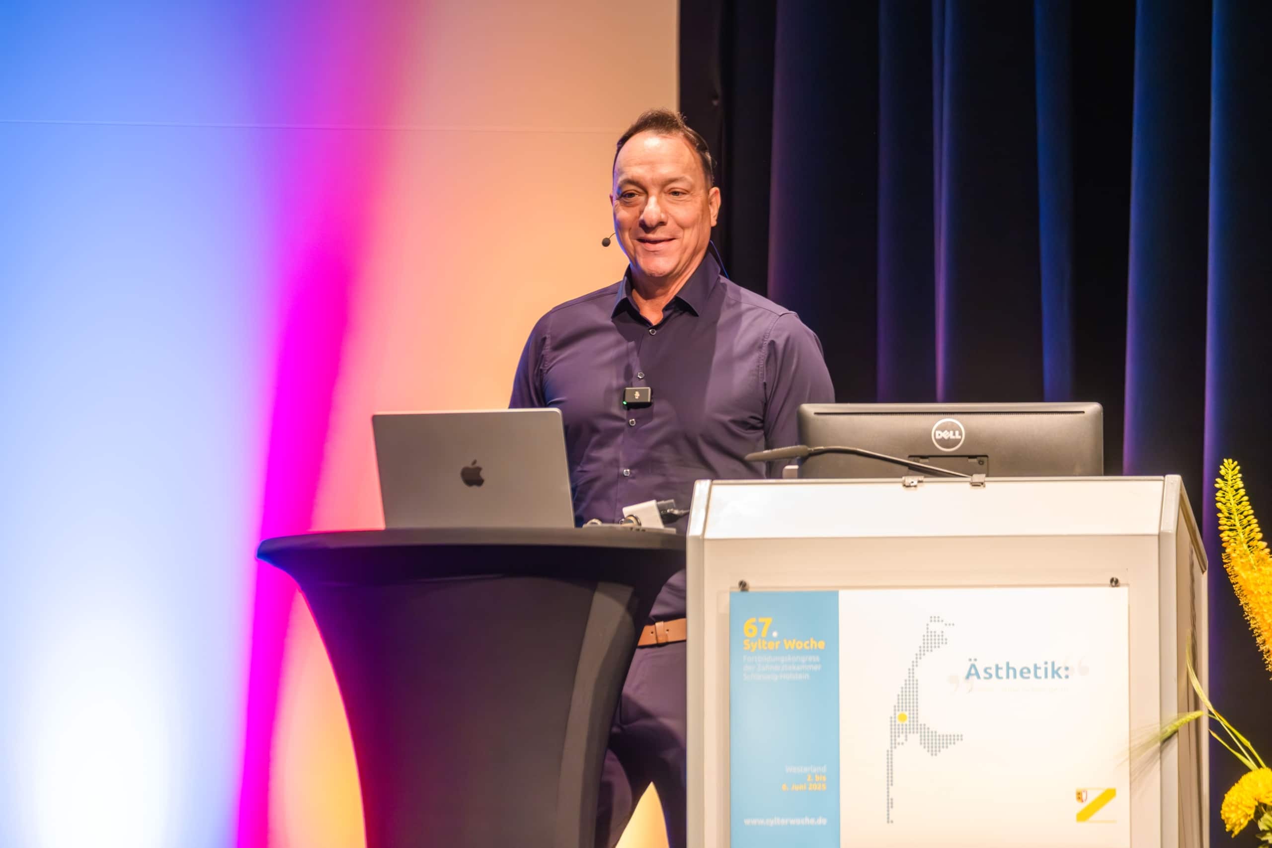 Ein Mann in einem dunklen Hemd steht an einem Podium mit einem Laptop und einem Mikrofon-Headset und hält lächelnd eine Präsentation. Im Hintergrund sind ein Lichtverlauf und ein Schild mit der Aufschrift 
