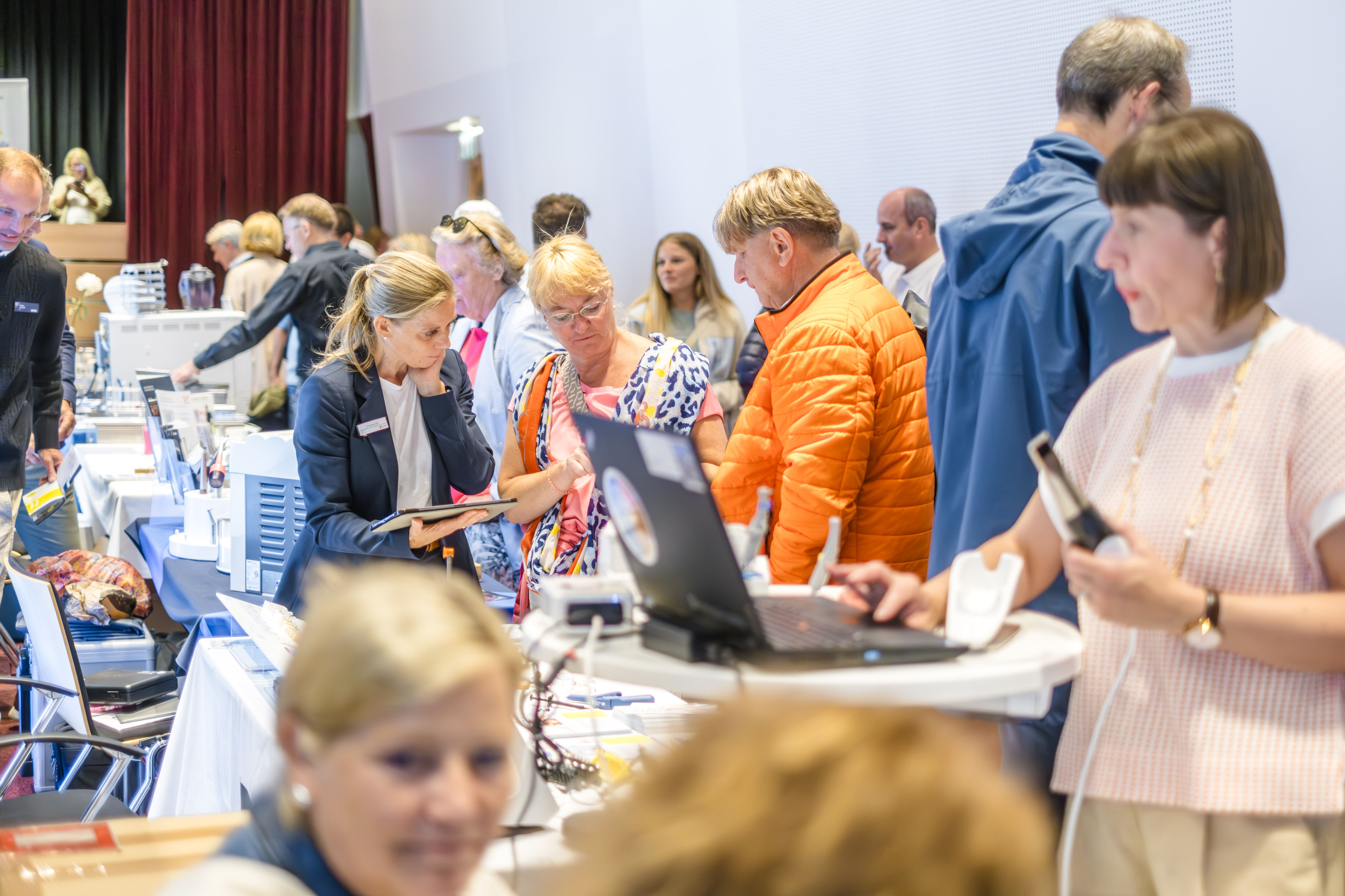Bei einer geschäftigen Veranstaltung in einer Halle diskutieren und prüfen Menschen Gegenstände an Tischen mit Elektronik und Papieren. Eine Frau im Vordergrund arbeitet an einem Laptop, während andere um sie herum ein Gespräch führen.