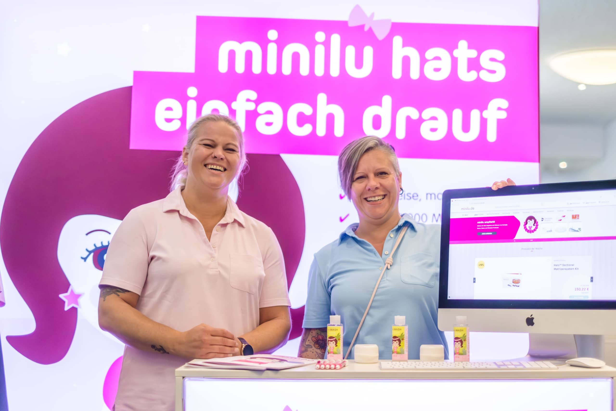 Zwei lächelnde Frauen stehen an einem Stand mit Produkten und einem Computermonitor vor einem leuchtend rosa-weißen Schild mit der Aufschrift 