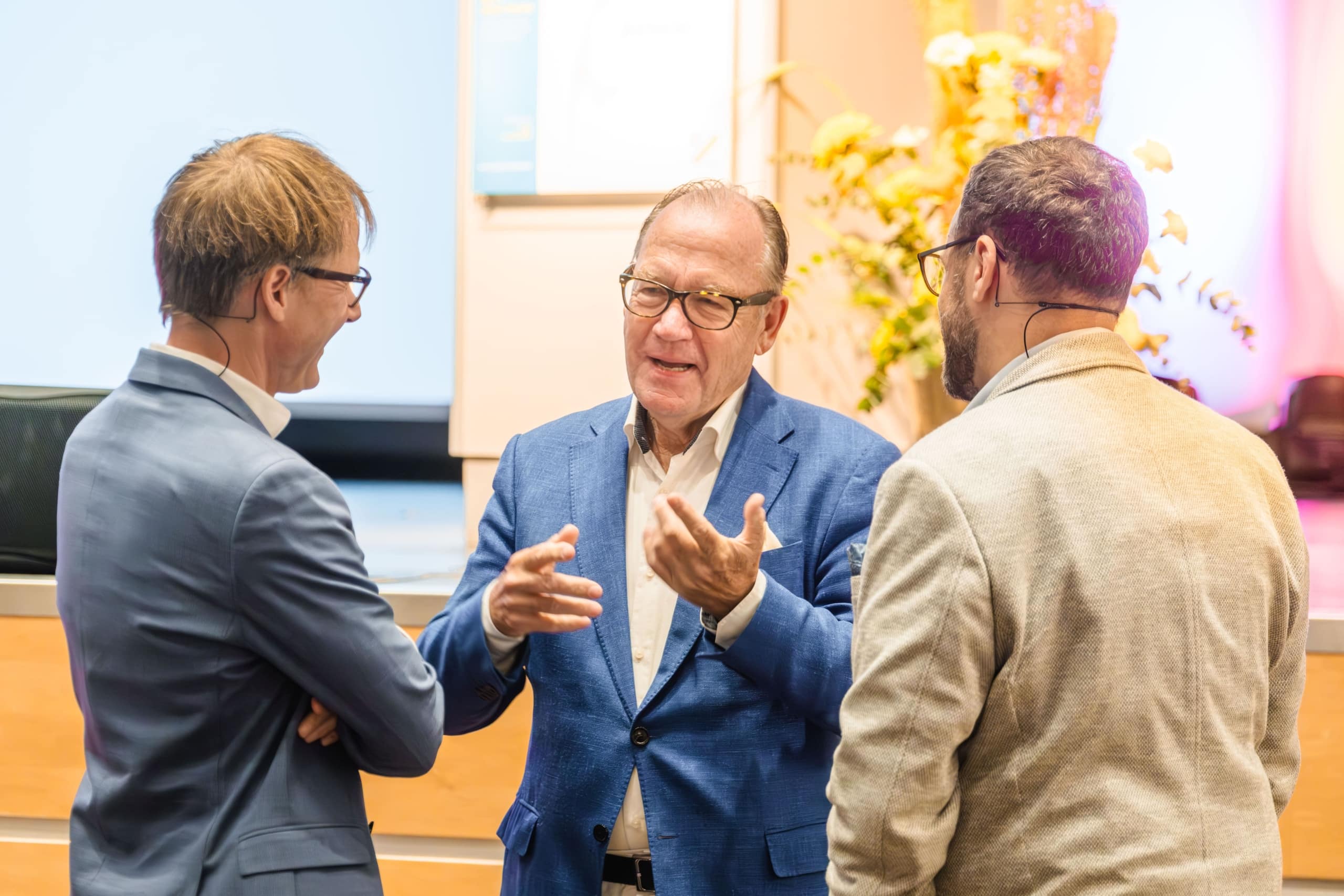 Drei Männer in Anzügen unterhalten sich in einem Innenraum in der Nähe einer Bühne, wobei einer gestikuliert, während er spricht. Im Hintergrund sind gelbe Blumen und sanfte Beleuchtung zu sehen.