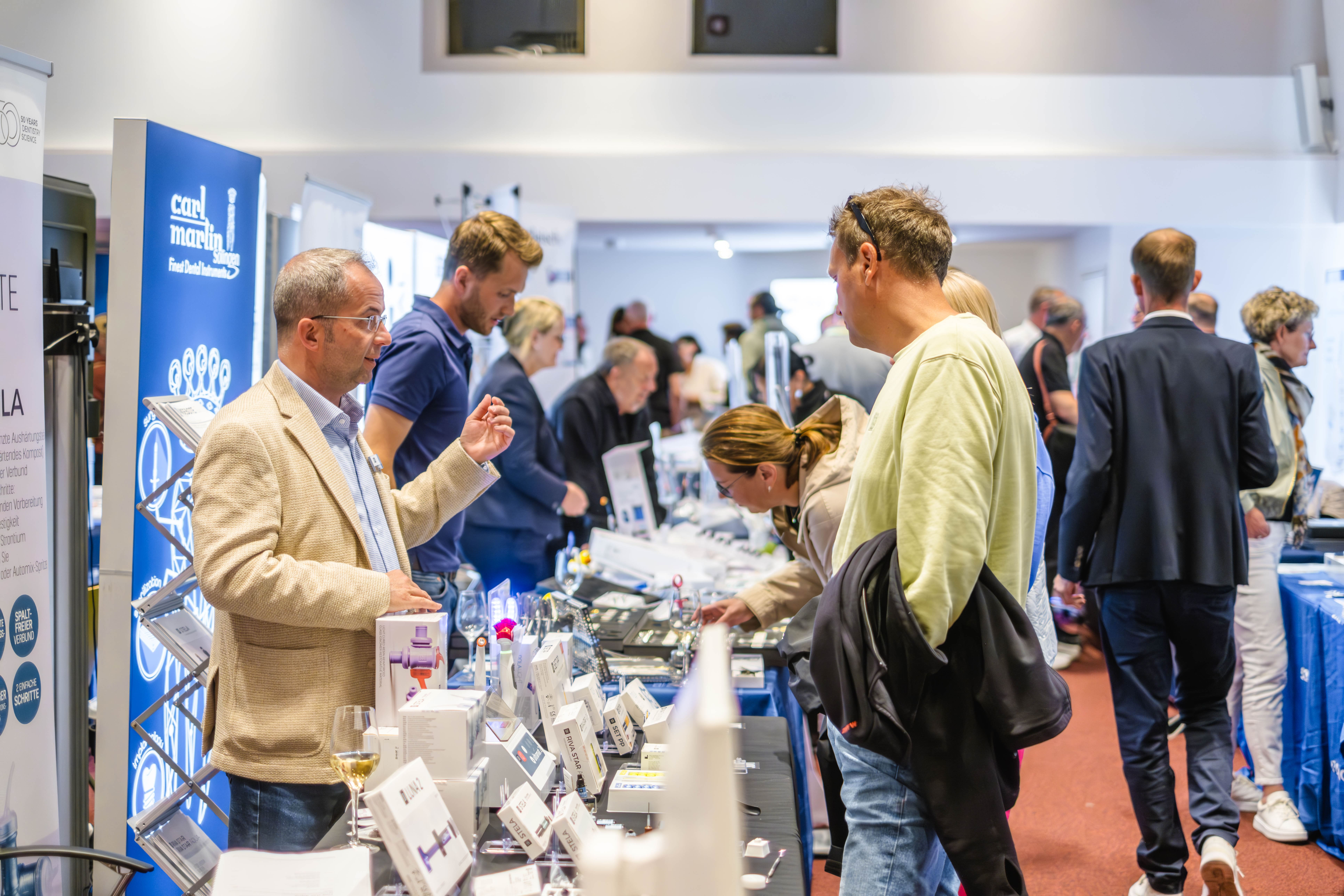 Auf einer belebten Messe oder Ausstellung mit Schautischen, Broschüren und elektronischen Geräten herrscht reger Austausch. Mehrere Teilnehmer sind in ein Gespräch vertieft oder sehen sich Produkte an. Die Atmosphäre ist professionell und lebhaft.