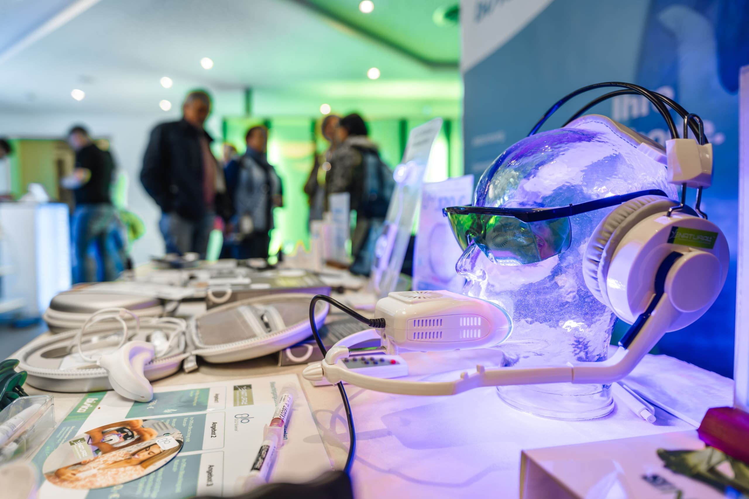 Ein Glaskopfmodell mit Kopfhörern und Sonnenbrille wird auf einem Tisch mit elektronischen Geräten und Broschüren ausgestellt, während im unscharfen Hintergrund bei einer Indoor-Veranstaltung Menschen stehen und interagieren.