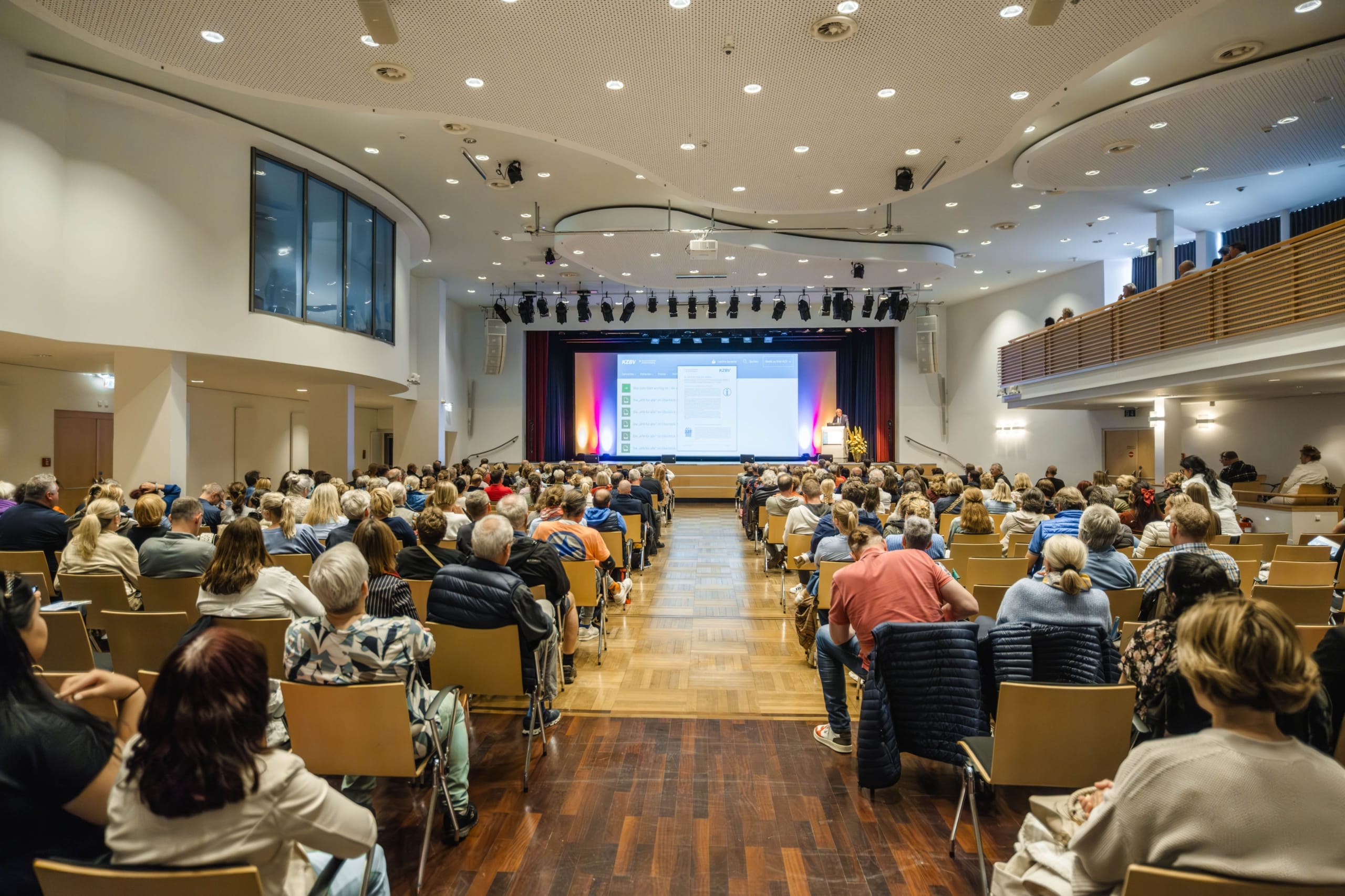 In einem geräumigen, modernen Auditorium sitzt ein großes Publikum in Reihen vor einer Bühne, auf die eine Präsentation auf eine Leinwand projiziert wird. Der Raum ist hell erleuchtet, und die Menschen beobachten aufmerksam den Redner.