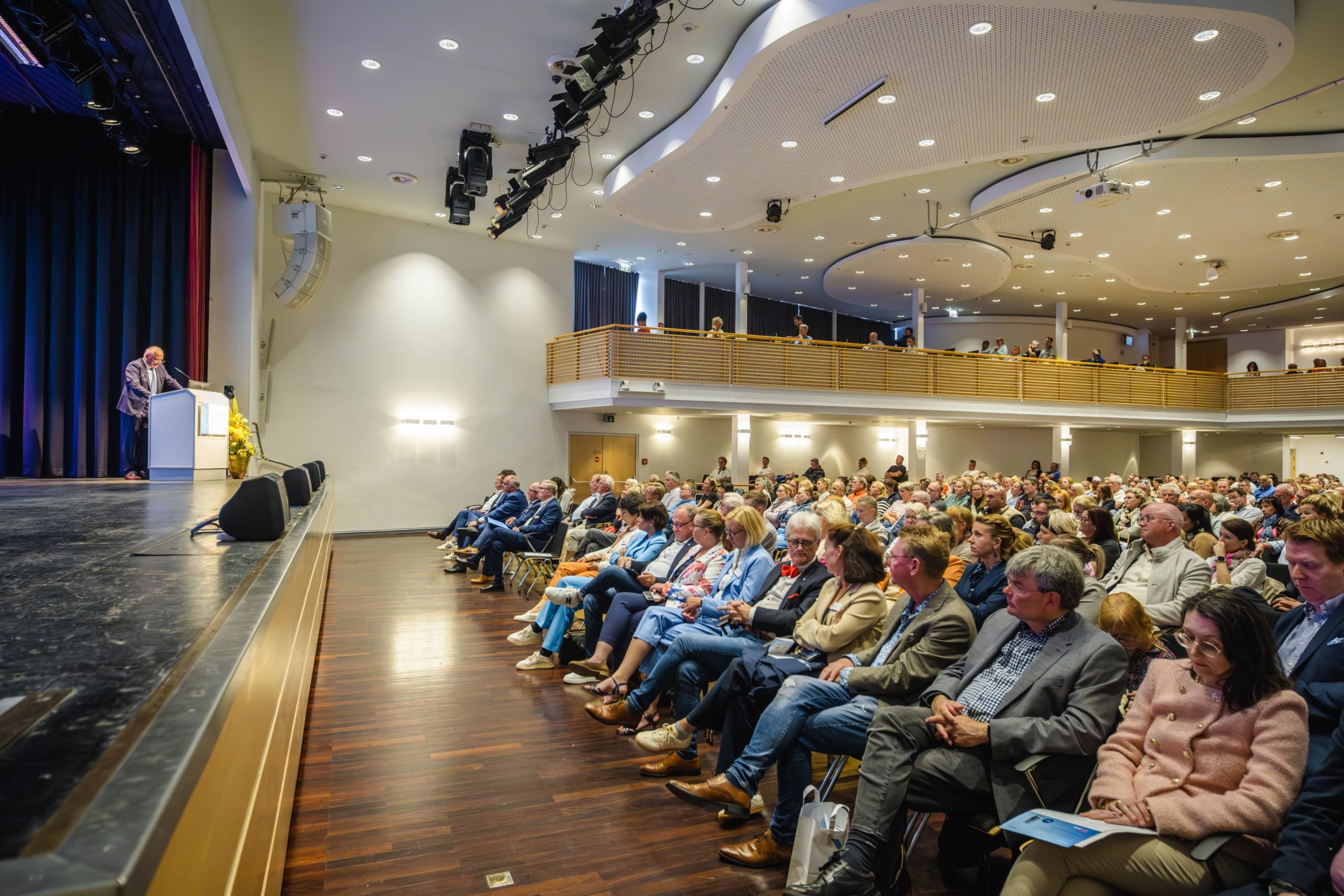 Ein Redner steht an einem Podium auf der Bühne und spricht zu einem großen, sitzenden Publikum in einem modernen Auditorium mit Balkonbestuhlung und heller Deckenbeleuchtung.