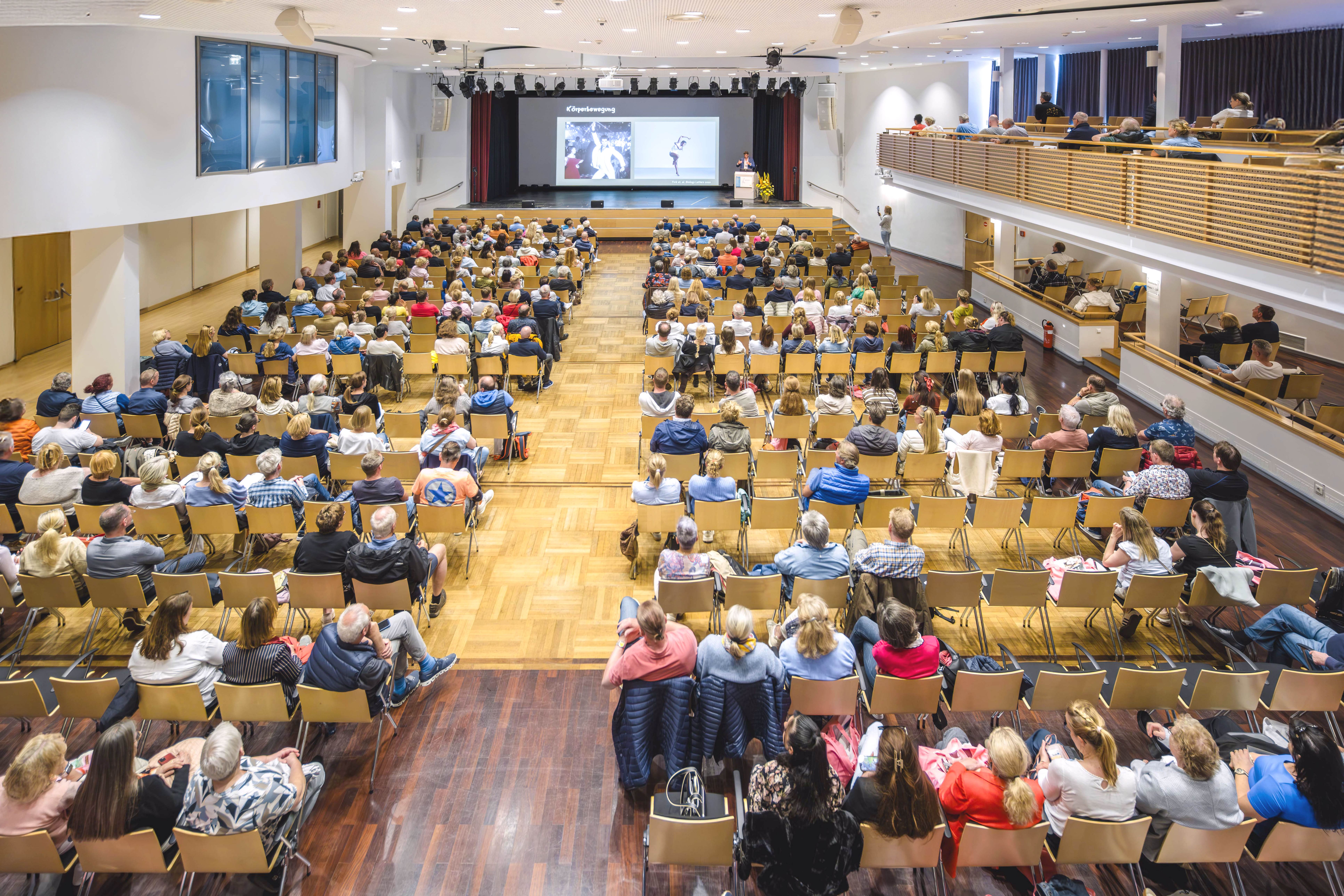 Ein großes Publikum sitzt in Stuhlreihen in einem geräumigen Auditorium und verfolgt eine Präsentation auf einer Bühne mit einer Leinwand, auf der Bilder gezeigt werden. Der Raum hat einen Holzboden und einen Balkon mit zusätzlichen Sitzgelegenheiten.