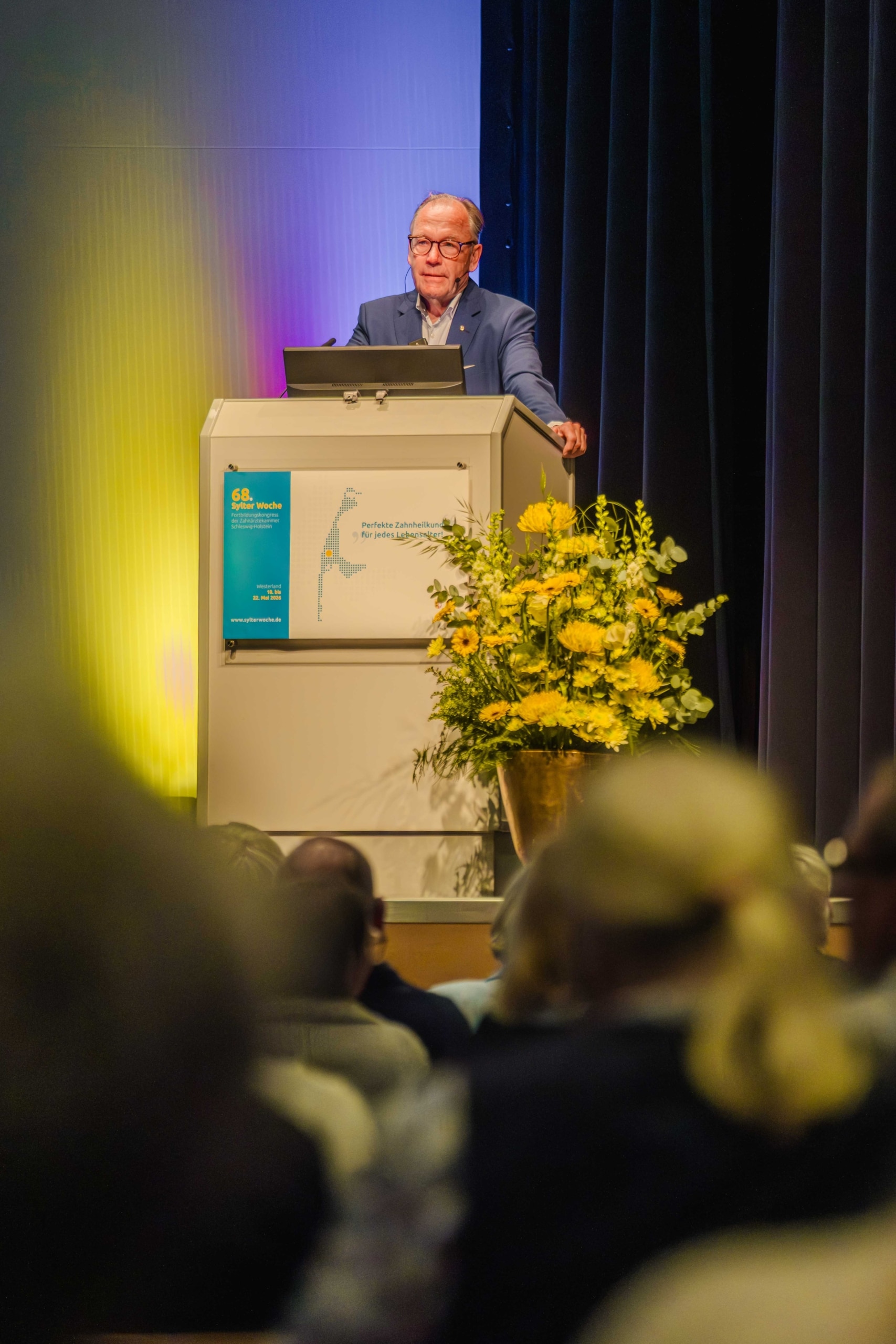 Ein älterer Mann im Anzug steht am Rednerpult und hält vor einem Publikum einen Vortrag. Neben dem Rednerpult stehen gelbe Blumen, und vorne ist ein blau-gelbes Veranstaltungsschild angebracht.