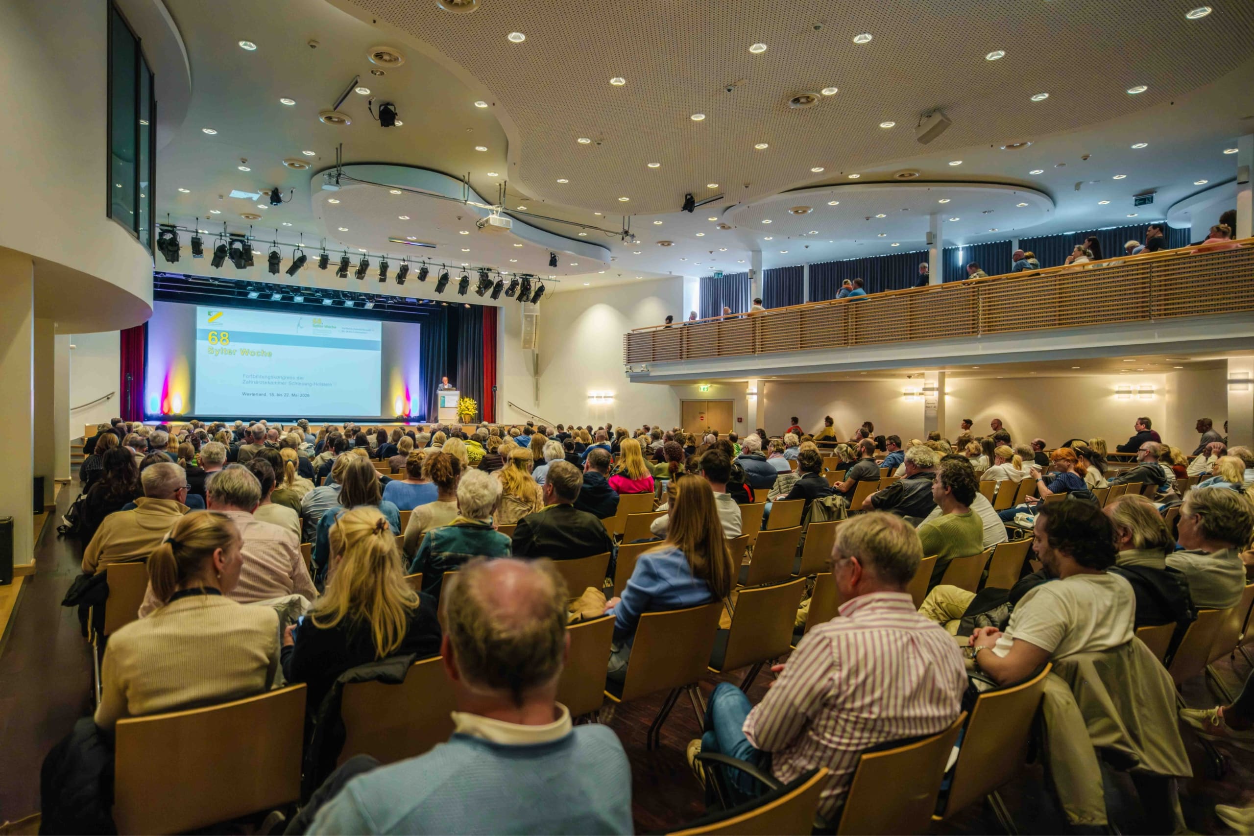 In einem modernen Auditorium sitzt ein großes Publikum vor einer Bühne, auf der ein Redner am Rednerpult steht, während eine Präsentation auf eine Leinwand projiziert wird. Der Saal ist gut beleuchtet und verfügt sowohl über Sitzplätze im Parkett als auch auf dem Rang.