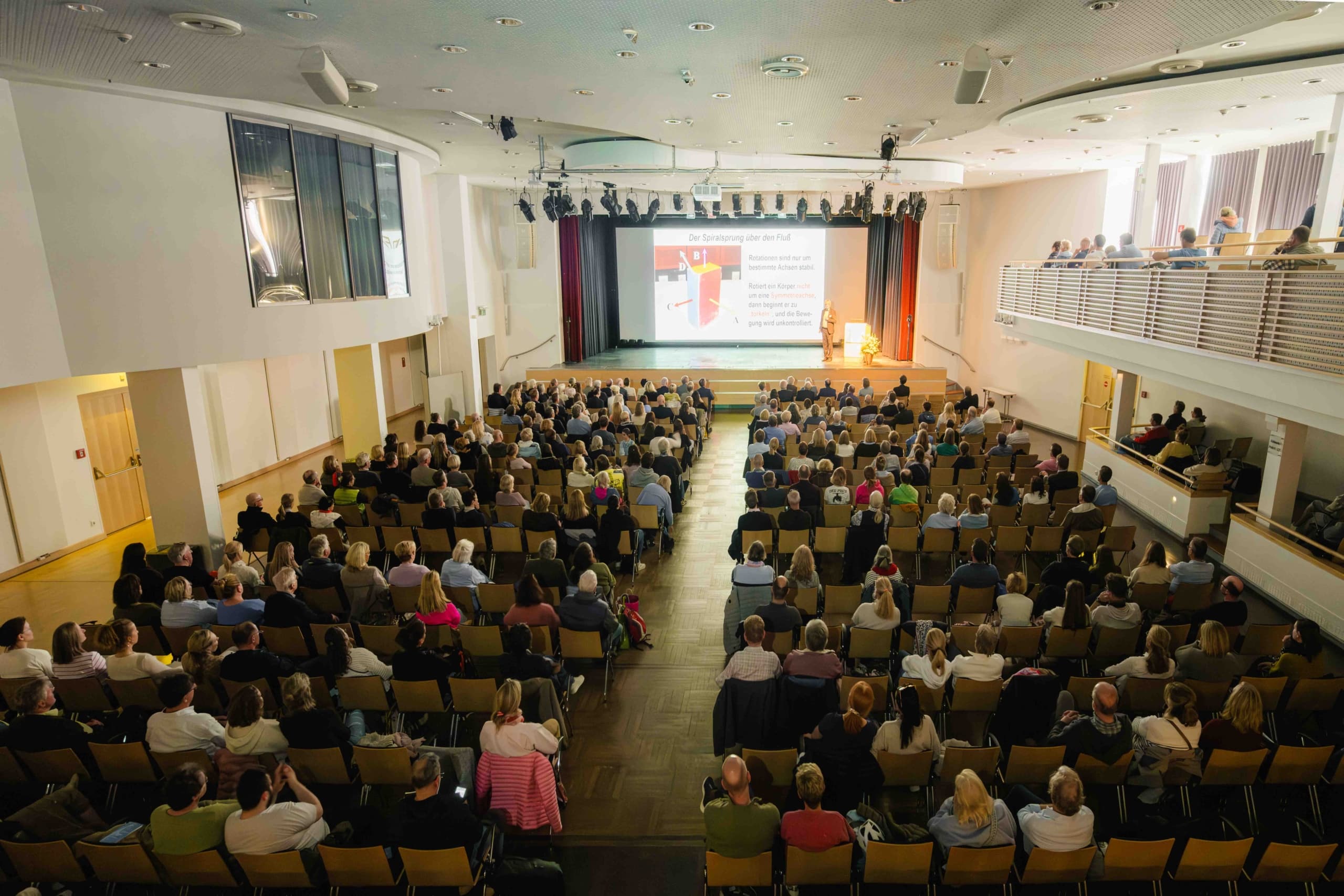 In einem hellen, modernen Saal sitzt ein großes Publikum in Reihen vor einer Bühne, auf der eine Präsentation auf eine Leinwand projiziert wird. Einige Personen sitzen rechts auf der Empore.