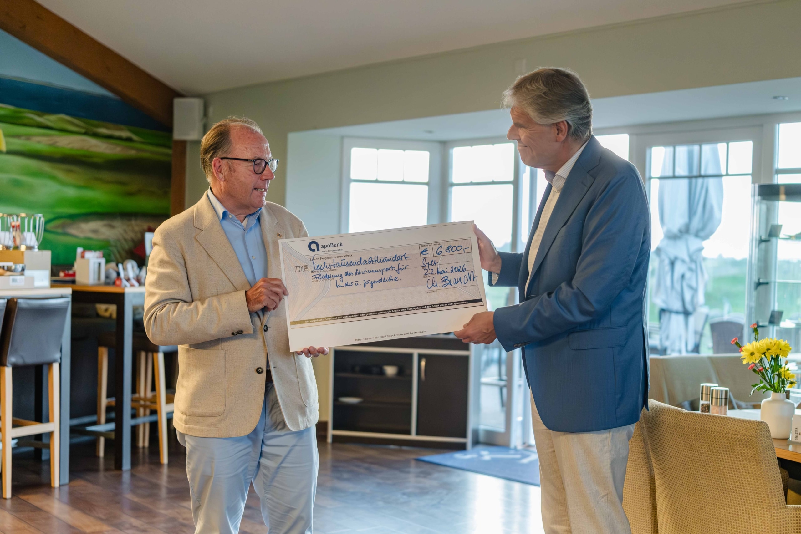 Zwei Männer in Anzügen stehen in einem Raum, während der eine dem anderen einen großen symbolischen Scheck über 6.000 € überreicht. Sie lächeln, im Hintergrund sind eine Bar und Fenster zu sehen, und auf einem Tisch in der Nähe steht eine Vase mit Blumen.