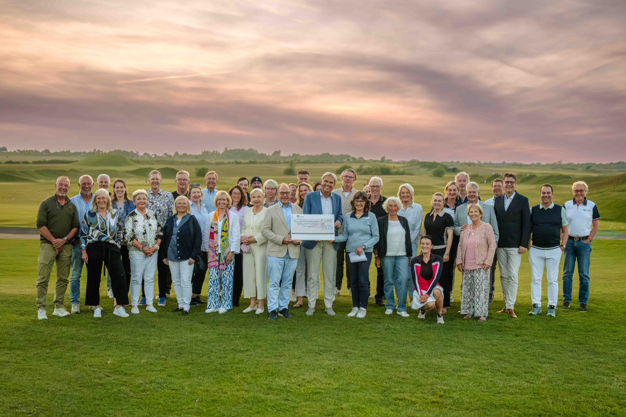 Eine große Gruppe von Menschen steht bei Sonnenuntergang auf einer Wiese und lächelt in die Kamera. Mehrere Personen in der Mitte halten einen großen symbolischen Scheck in den Händen. Im Hintergrund sind sanfte Hügel und ein farbenprächtiger Himmel zu sehen.