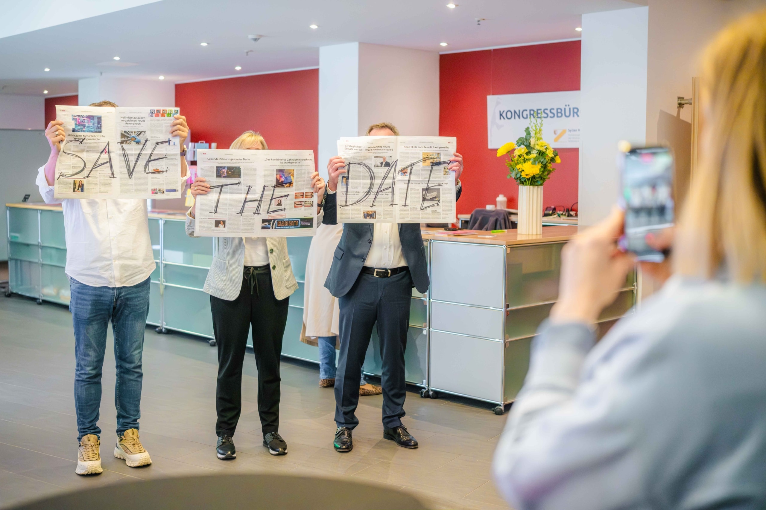 Drei Personen stehen in einem Raum und halten Zeitungen in der Hand, auf denen „SAVE THE DATE“ steht, während eine weitere Person sie mit einem Smartphone fotografiert. Der Schauplatz scheint ein modernes Büro oder ein Veranstaltungsraum zu sein.