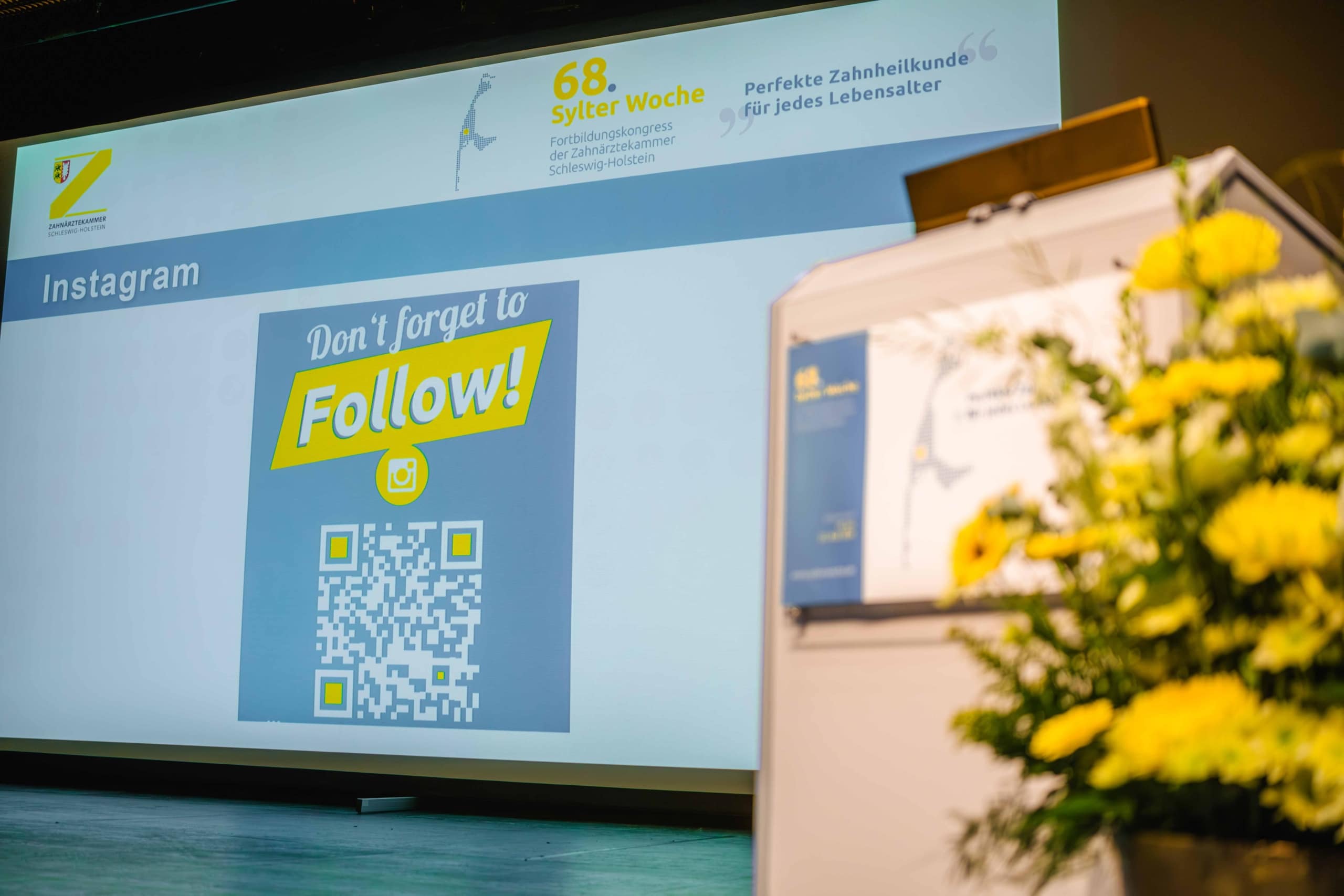 Auf einer Präsentationsfolie ist ein QR-Code mit dem Text „Vergiss nicht, uns auf Instagram zu folgen!“ zu sehen. Im Vordergrund sind gelbe Blumen und ein Podium zu sehen. Auf einem Banner darüber steht „68. Sylter Woche“ sowie Informationen zur Zahnpflege.