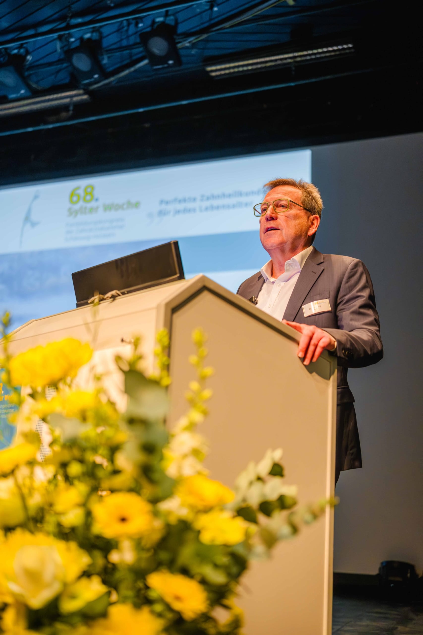 Ein Mann im Anzug steht an einem Rednerpult und hält einen Vortrag; im Vordergrund sind gelbe Blumen zu sehen, im Hintergrund eine projizierte Folie mit deutschem Text.