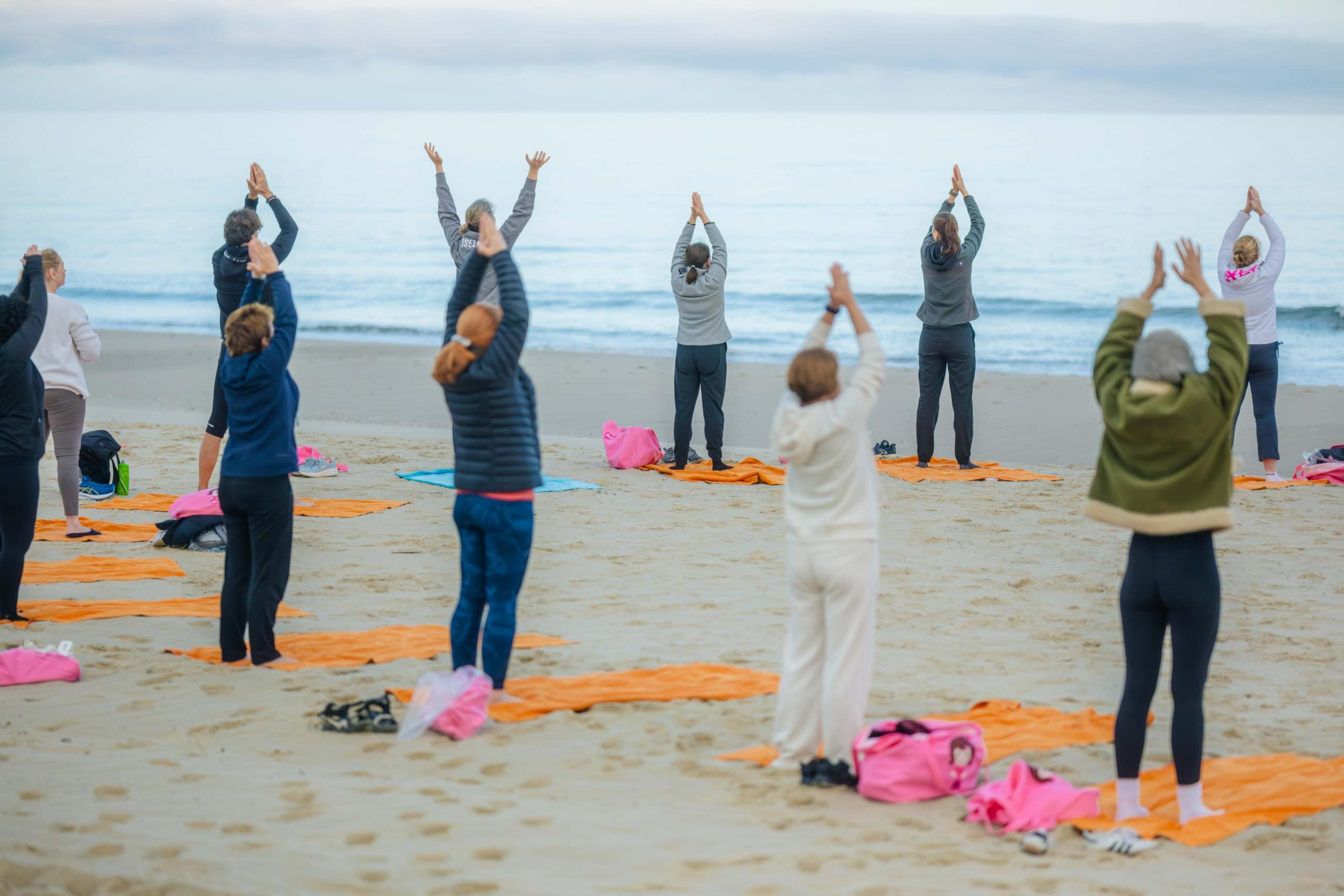 Eine Gruppe von Menschen praktiziert Yoga auf orangefarbenen Matten an einem Sandstrand; alle stehen mit erhobenen Armen da. Im Hintergrund sind das Meer und der bewölkte Himmel zu sehen. Die Teilnehmer tragen Jacken und warme Kleidung.
