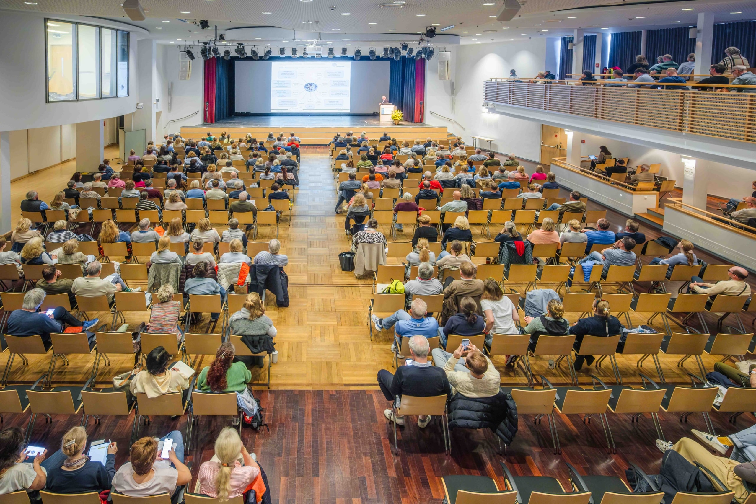 In einem geräumigen, gut beleuchteten Saal sitzt ein großes Publikum in Stuhlreihen mit Blick auf die Bühne. Eine Person steht am Rednerpult und hält einen Vortrag, während hinter ihr auf einer großen Leinwand Bildmaterial gezeigt wird.