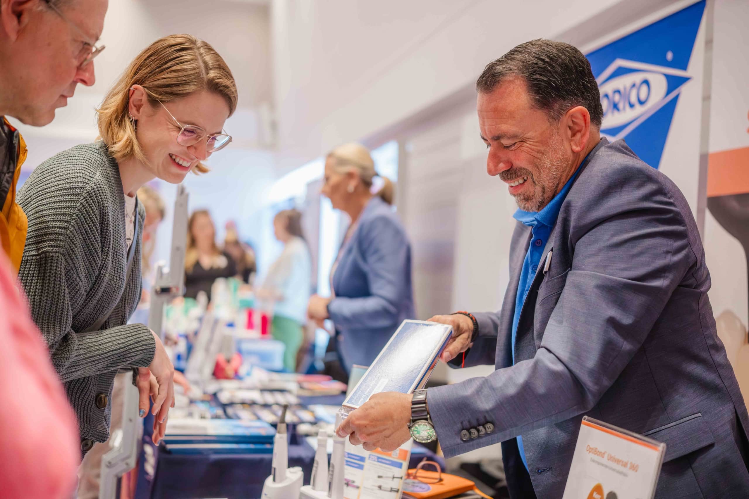 Ein lächelnder Mann im Anzug präsentiert einer Frau an einem Messestand in einer Halle ein Produkt. Im Hintergrund sind weitere Besucher und Ausstellungsstücke zu sehen. Die Atmosphäre wirkt freundlich und einladend.