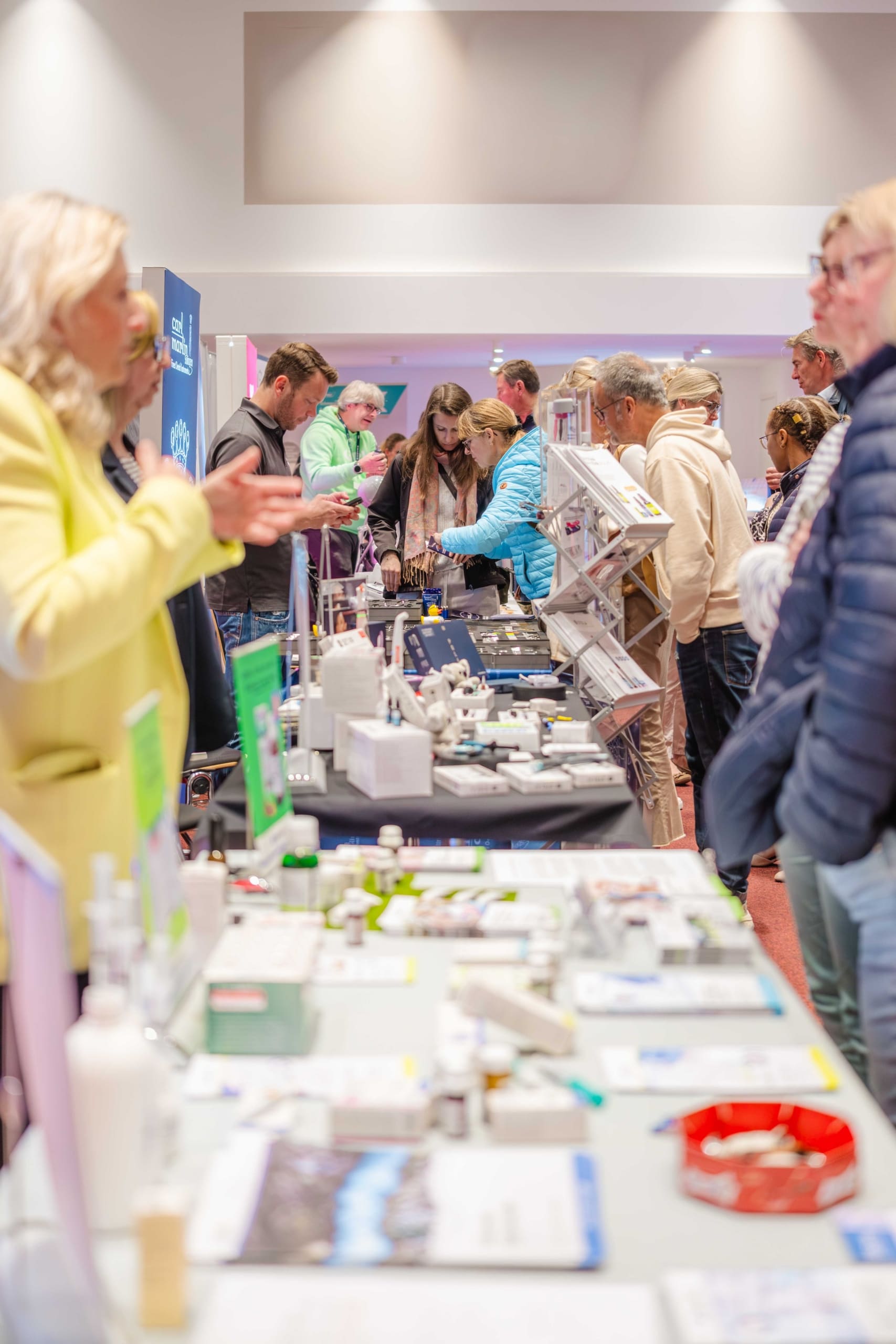 Eine belebte Messe oder Konferenz in einem Innenraum, bei der sich die Besucher an Tischen austauschen, auf denen unter heller Beleuchtung verschiedene Produkte, Broschüren und Informationsmaterialien ausgestellt sind.