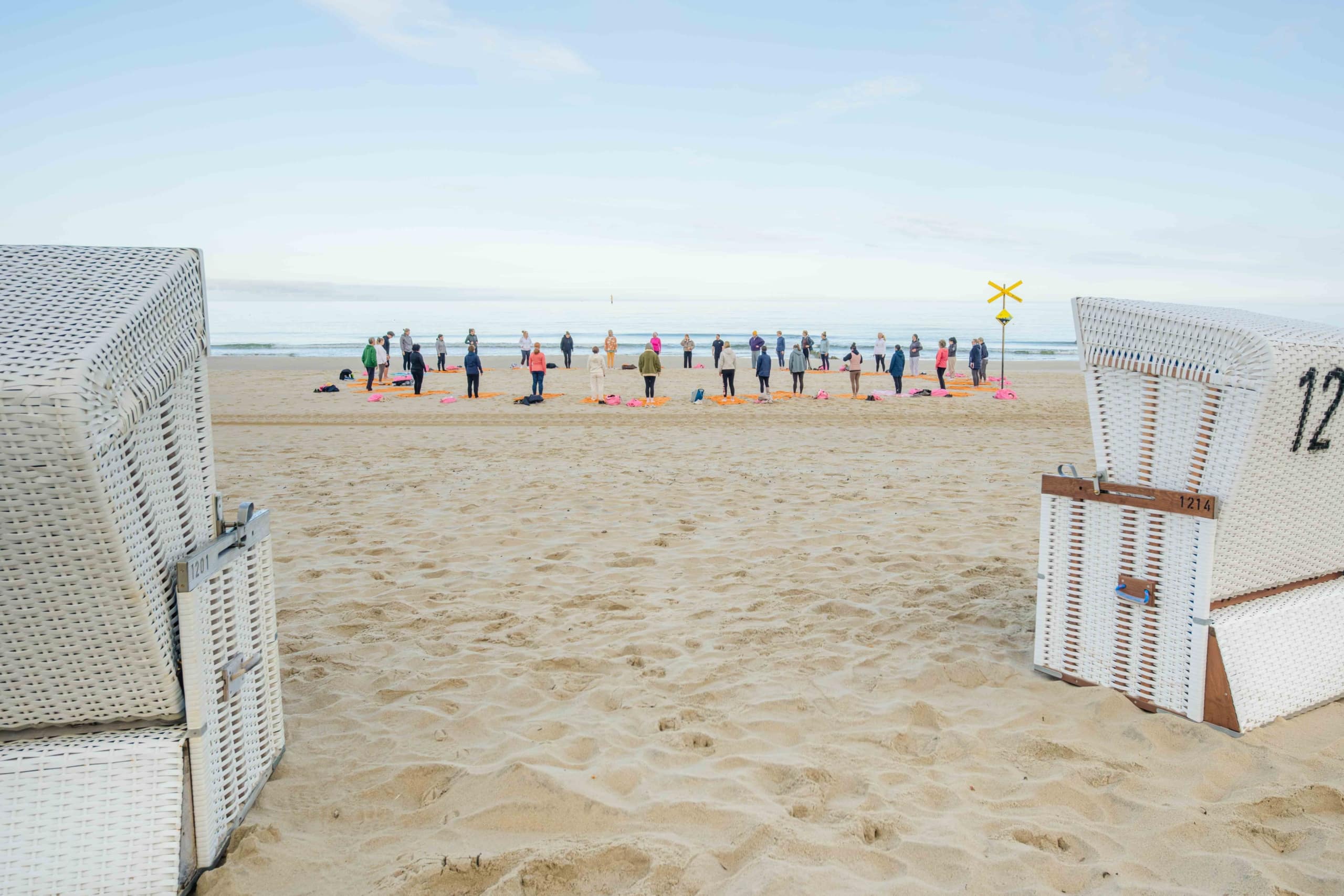 Eine Gruppe von Menschen steht in einer Reihe an einem Sandstrand nahe der Wasserlinie und macht Yoga oder Dehnübungen. Weiße Strandkörbe aus Korbgeflecht rahmen den Blick im Vordergrund ein. Der Himmel ist klar und ruhig.