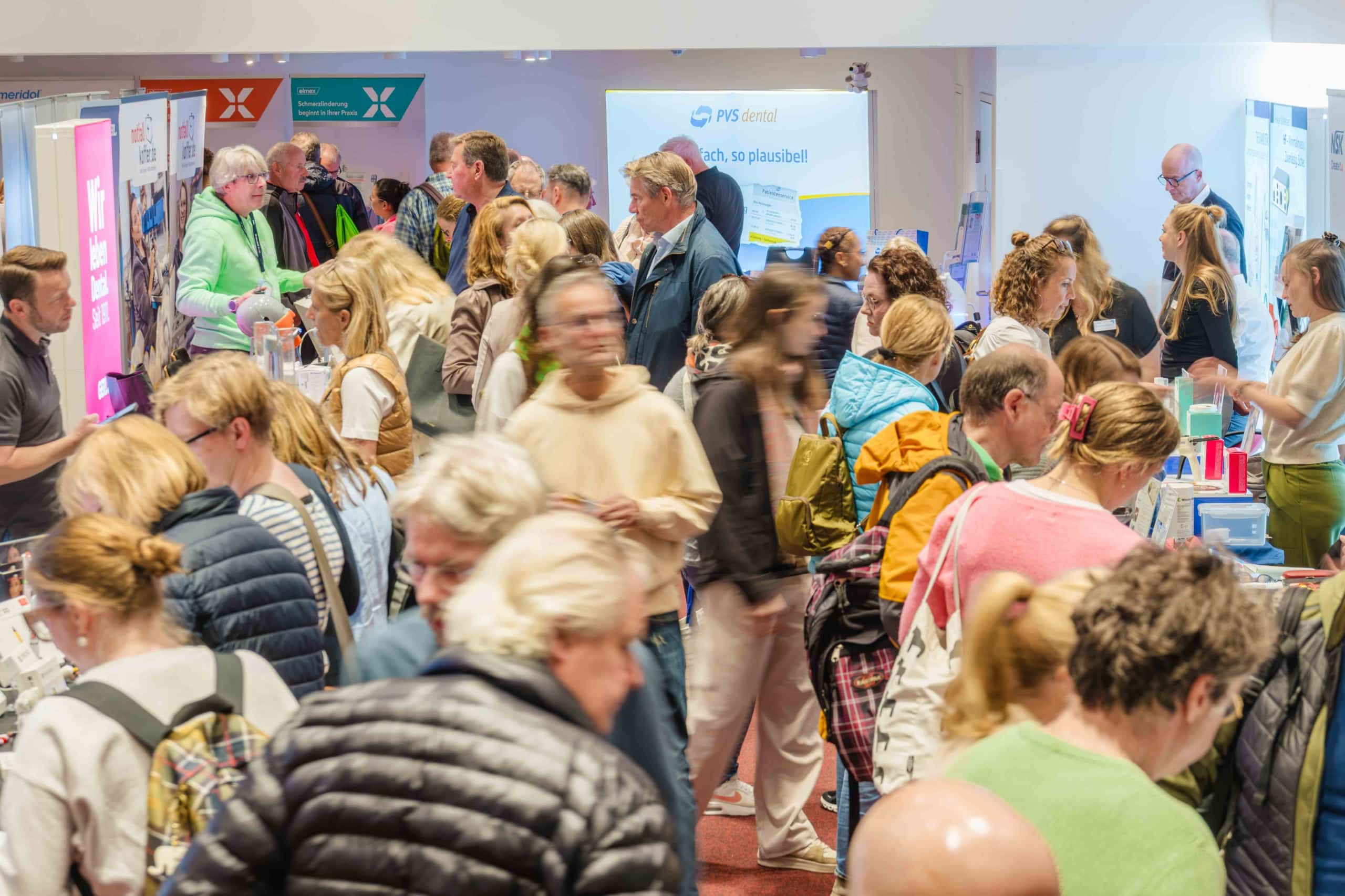 Eine gut besuchte Indoor-Veranstaltung, bei der sich viele Menschen an verschiedenen Ständen unterhalten, einige Broschüren in der Hand halten oder mit Ausstellern sprechen – in einer hell beleuchteten Halle, in der Banner und Werbematerialien zu sehen sind.