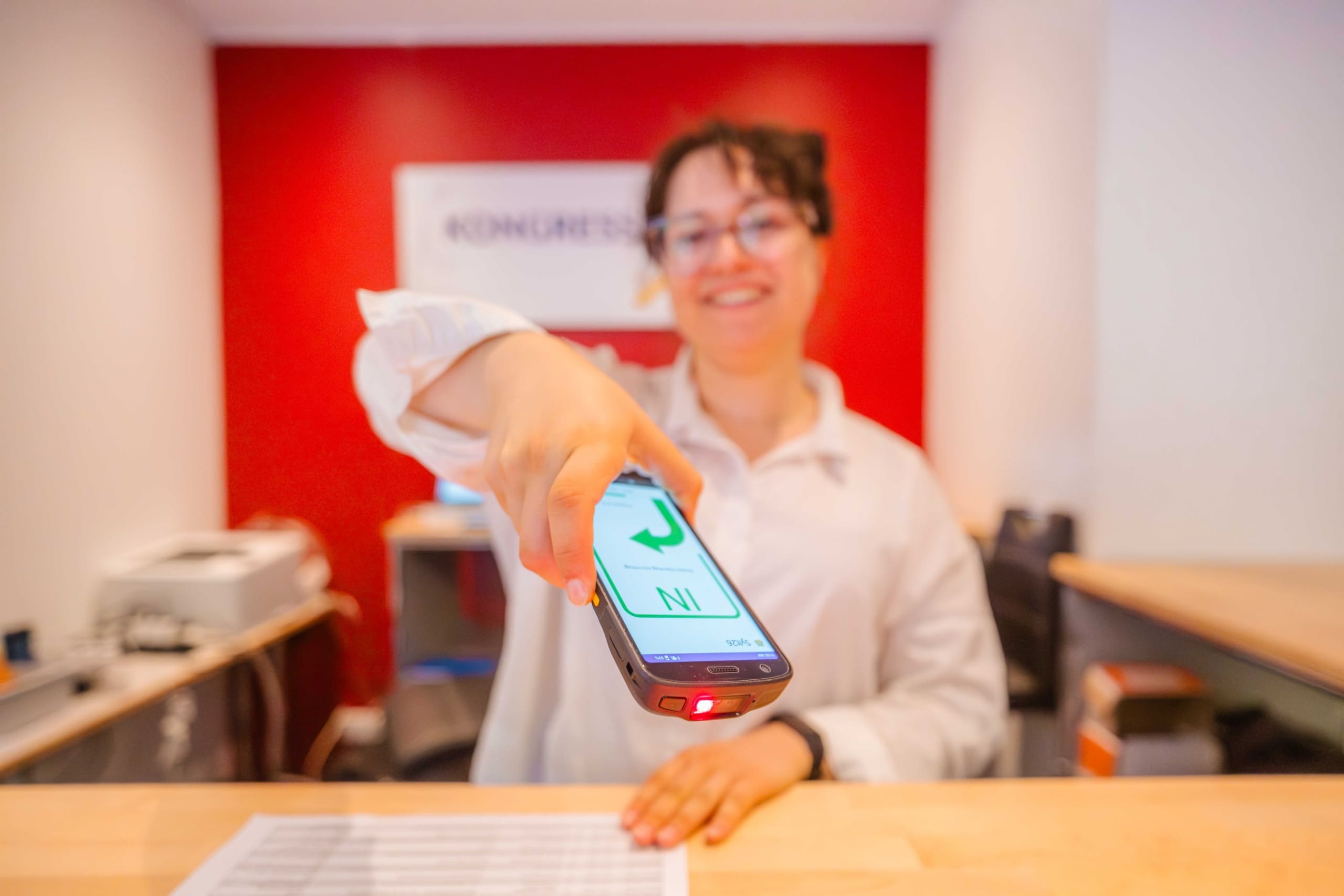 Eine lächelnde Rezeptionistin hält einen Hand-Barcodescanner mit einem grünen Häkchen auf dem Bildschirm in die Hand und steht an einem Empfangstresen vor einer roten Wand.