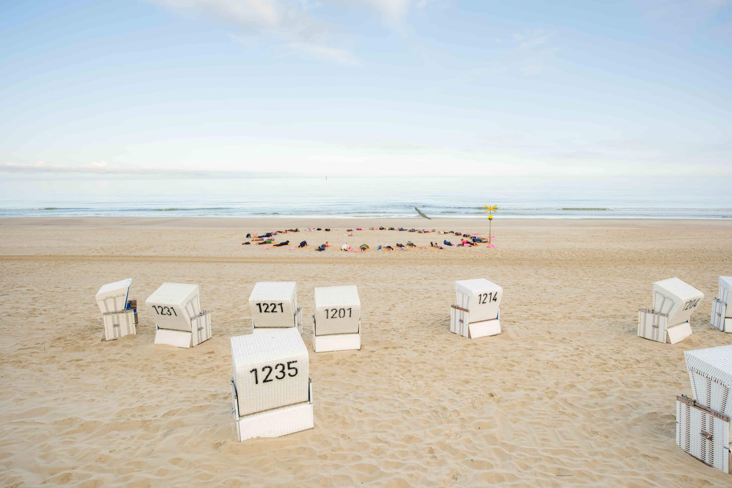 Ein Sandstrand mit weißen, nummerierten Liegestühlen im Vordergrund und einer Gruppe von Menschen, die auf Handtüchern in Ufernähe unter einem hellblauen Himmel liegen.