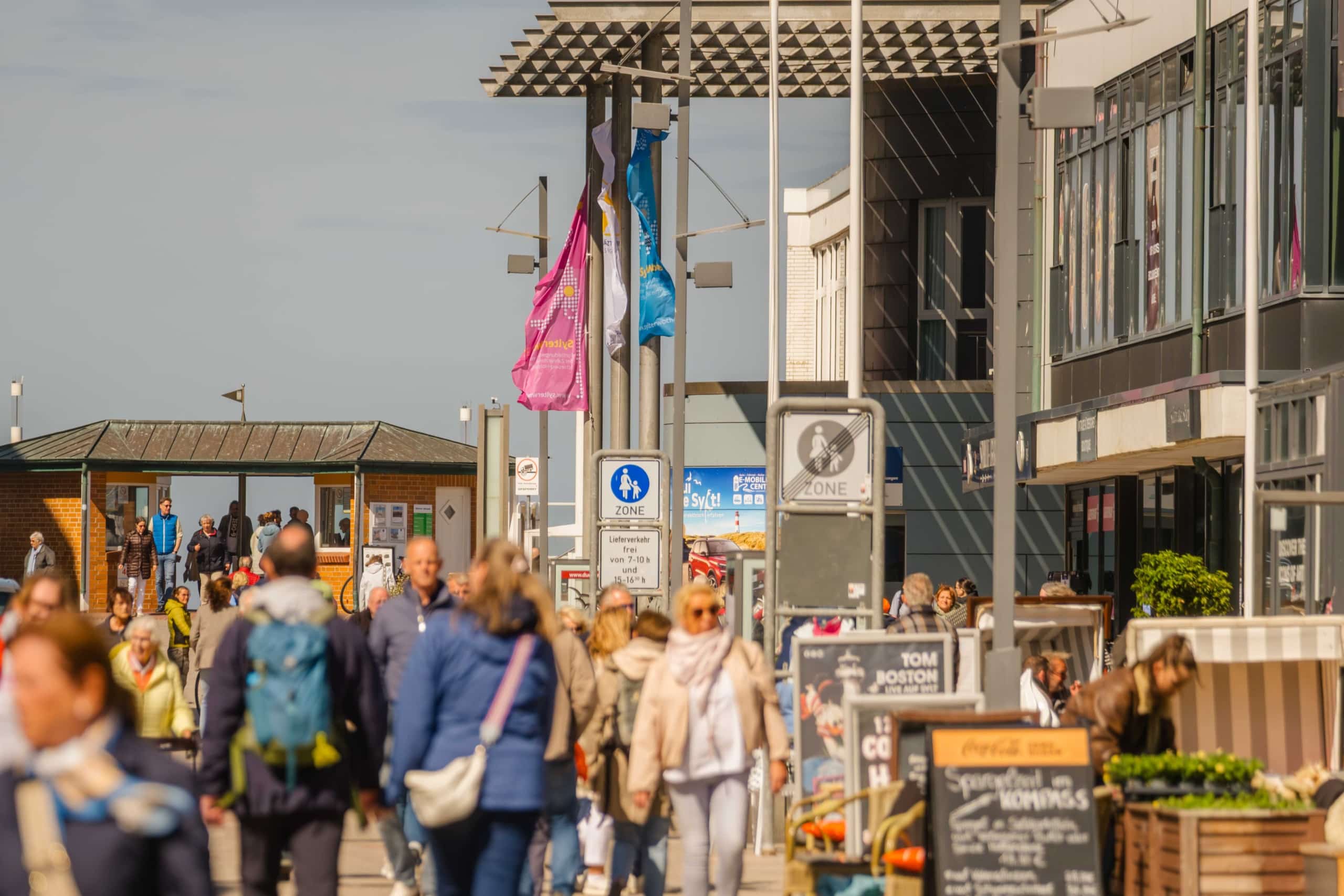 Eine belebte Fußgängerzone mit Passanten, bunten Fahnen, Ladenfronten und Sitzgelegenheiten im Freien an einem sonnigen Tag. Entlang des belebten Gehwegs sind Schilder und Speisekarten zu sehen.