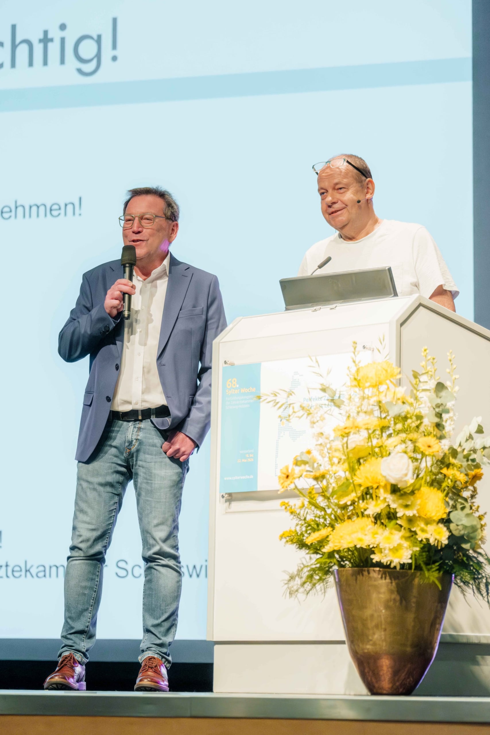 Zwei Männer stehen auf einer Bühne; einer spricht in ein Mikrofon, während der andere mit einem Laptop hinter einem Rednerpult steht. Ein großer Strauß gelber Blumen schmückt die Vorderseite des Rednerpults. Im Hintergrund ist eine Leinwand mit Text zu sehen.