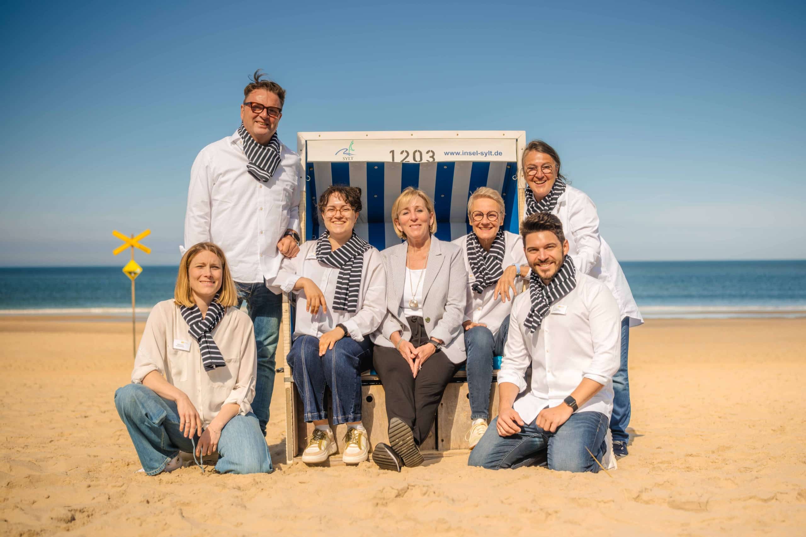 Sieben Erwachsene posieren lächelnd an einem Sandstrand vor dem Meer; sie tragen weiße Hemden und dazu passende gestreifte Schals. Eine Person sitzt auf einem großen Strandstuhl, während die anderen um sie herum stehen oder knien. Strahlendes, sonniges Wetter.