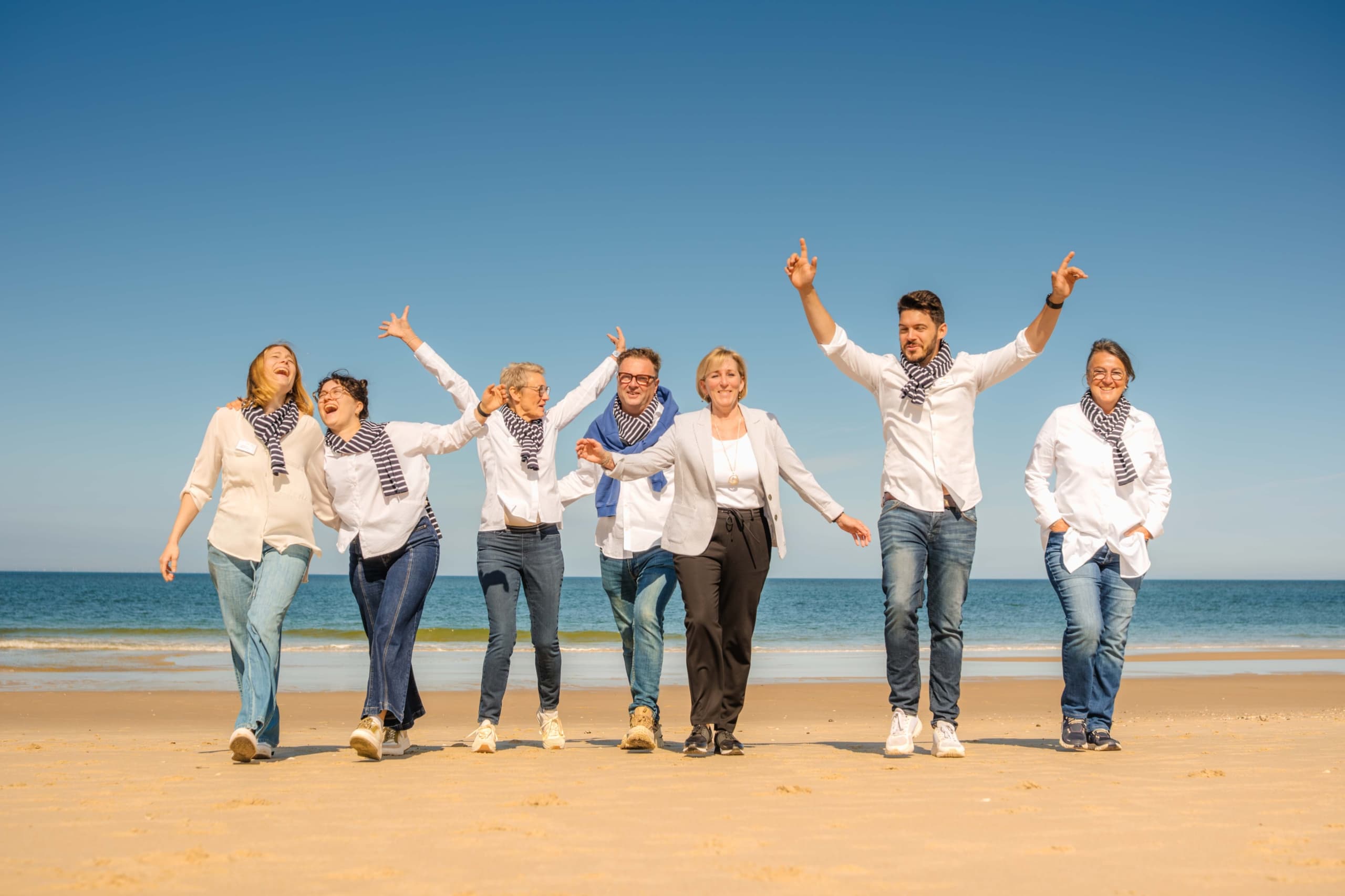 Eine Gruppe von sieben Personen, die weiße Oberteile, blaue Jeans und gestreifte Schals tragen, spaziert lachend gemeinsam an einem Sandstrand unter strahlend blauem Himmel und genießt den sonnigen Tag.