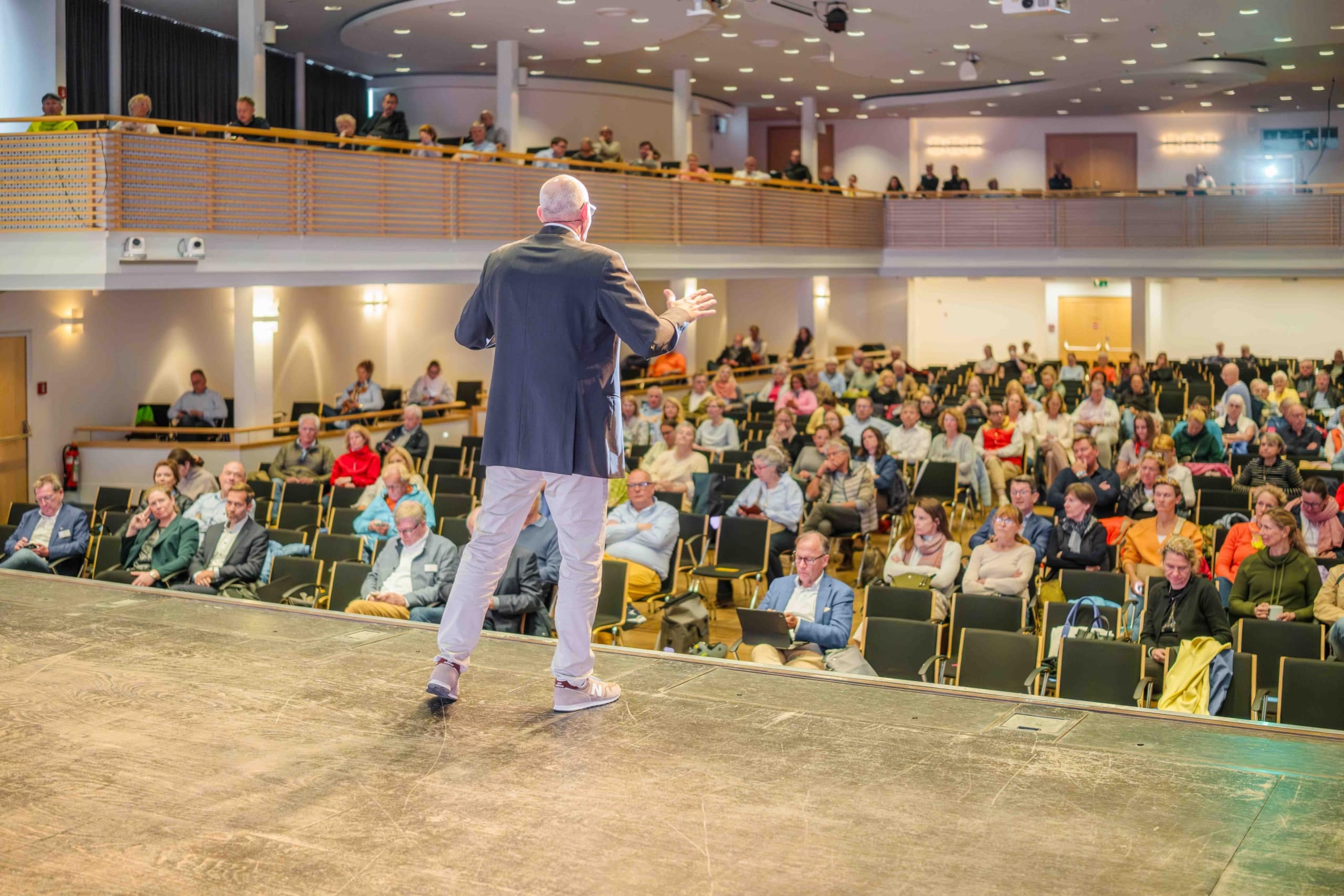 Ein Mann im Anzug steht auf der Bühne und blickt auf das Publikum, das in Reihen in einem großen, hellen Saal sitzt. Das Publikum wirkt aufmerksam, und es sitzen sowohl im Parkett als auch auf dem Rang Menschen.