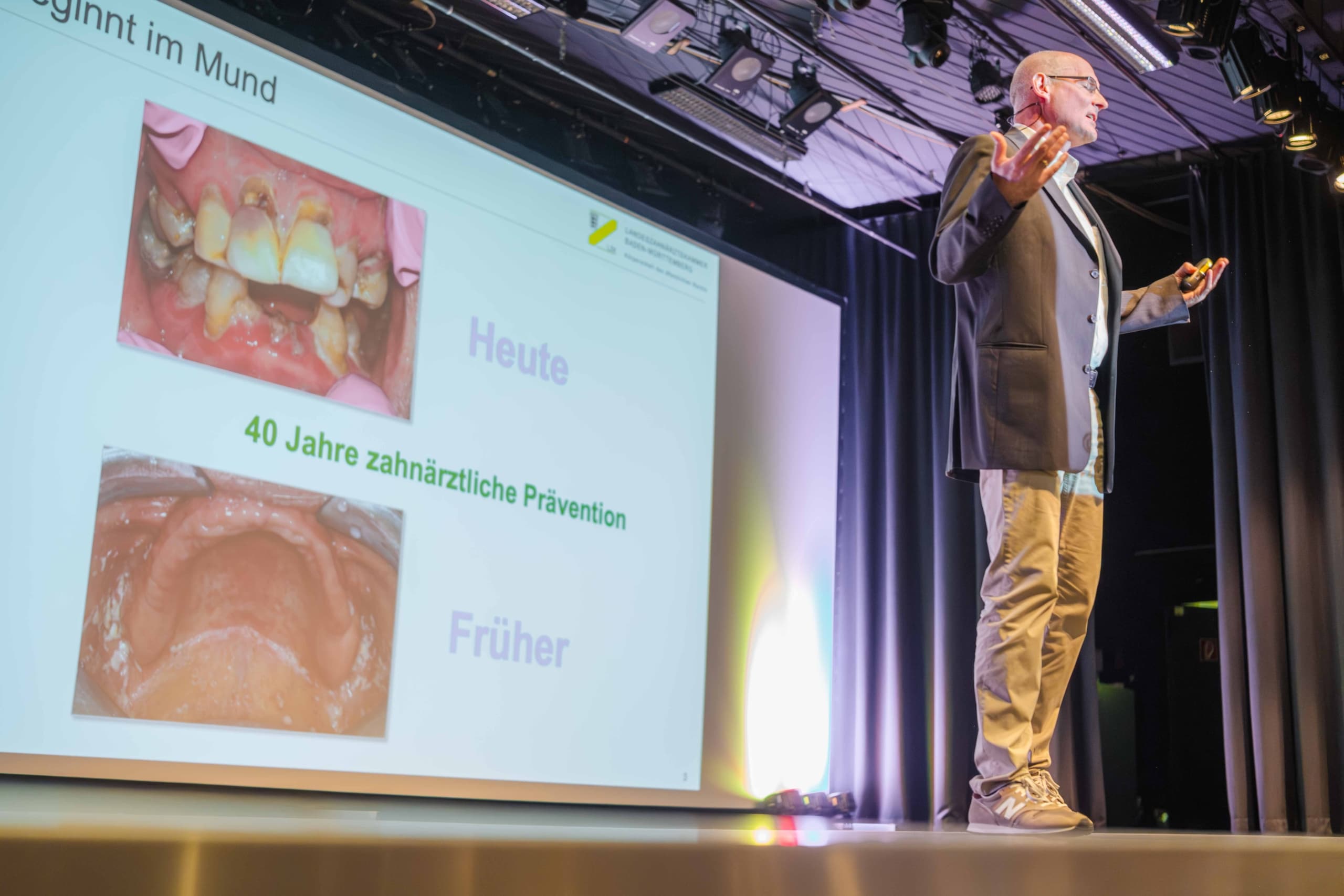 Ein Mann hält auf der Bühne einen Vortrag und zeigt dabei eine Folie mit Bildern zum Thema Mundgesundheit, die mit „Heute“ und „Früher“ beschriftet sind, um 40 Jahre zahnmedizinische Prävention in Deutschland zu veranschaulichen.