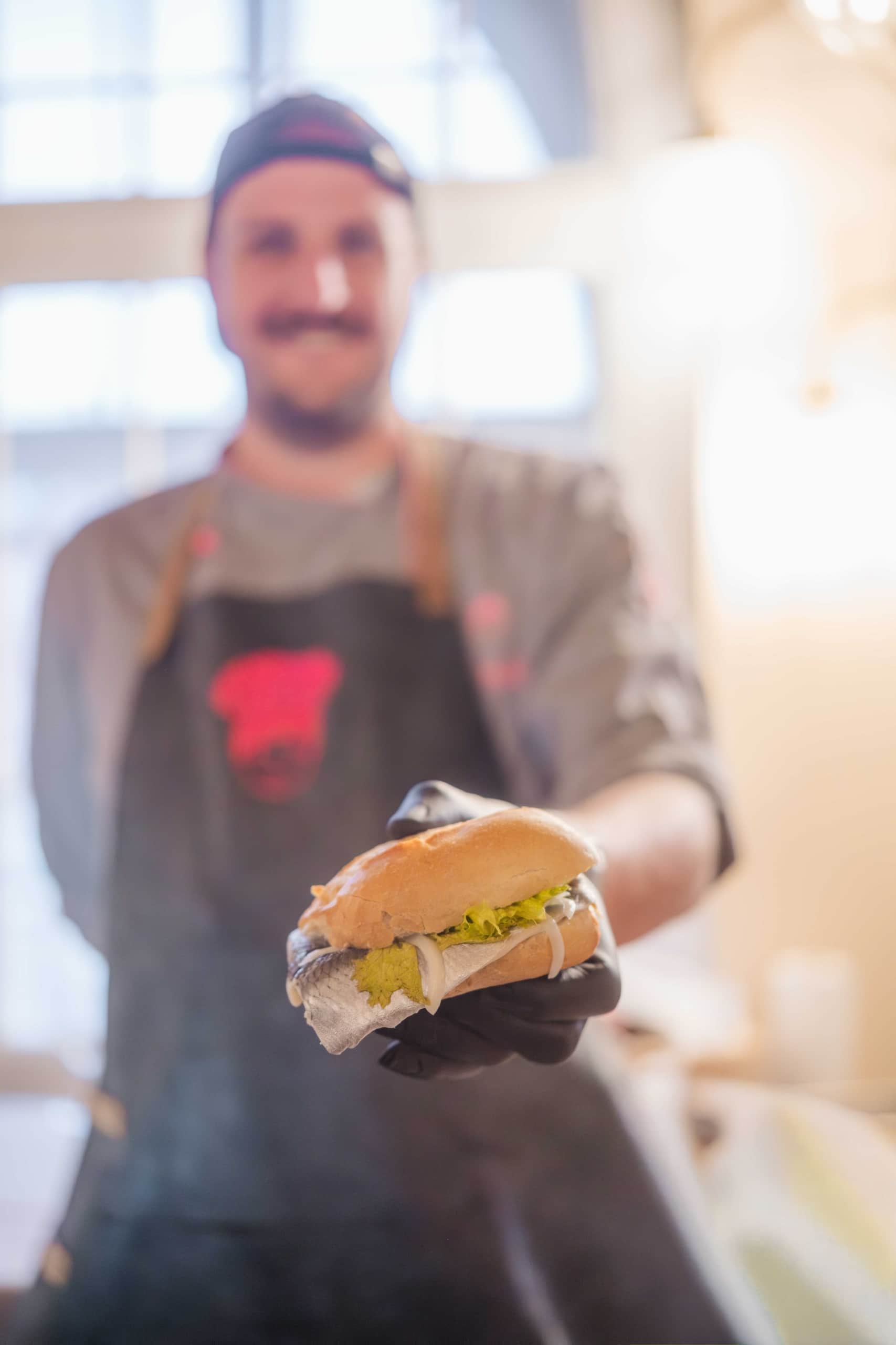 Ein Koch in einer dunklen Schürze und einer Kochmütze hält ein Sandwich mit Salat, Käse und Fleisch hoch und lächelt in einer warm beleuchteten Küche mit unscharfem Hintergrund.