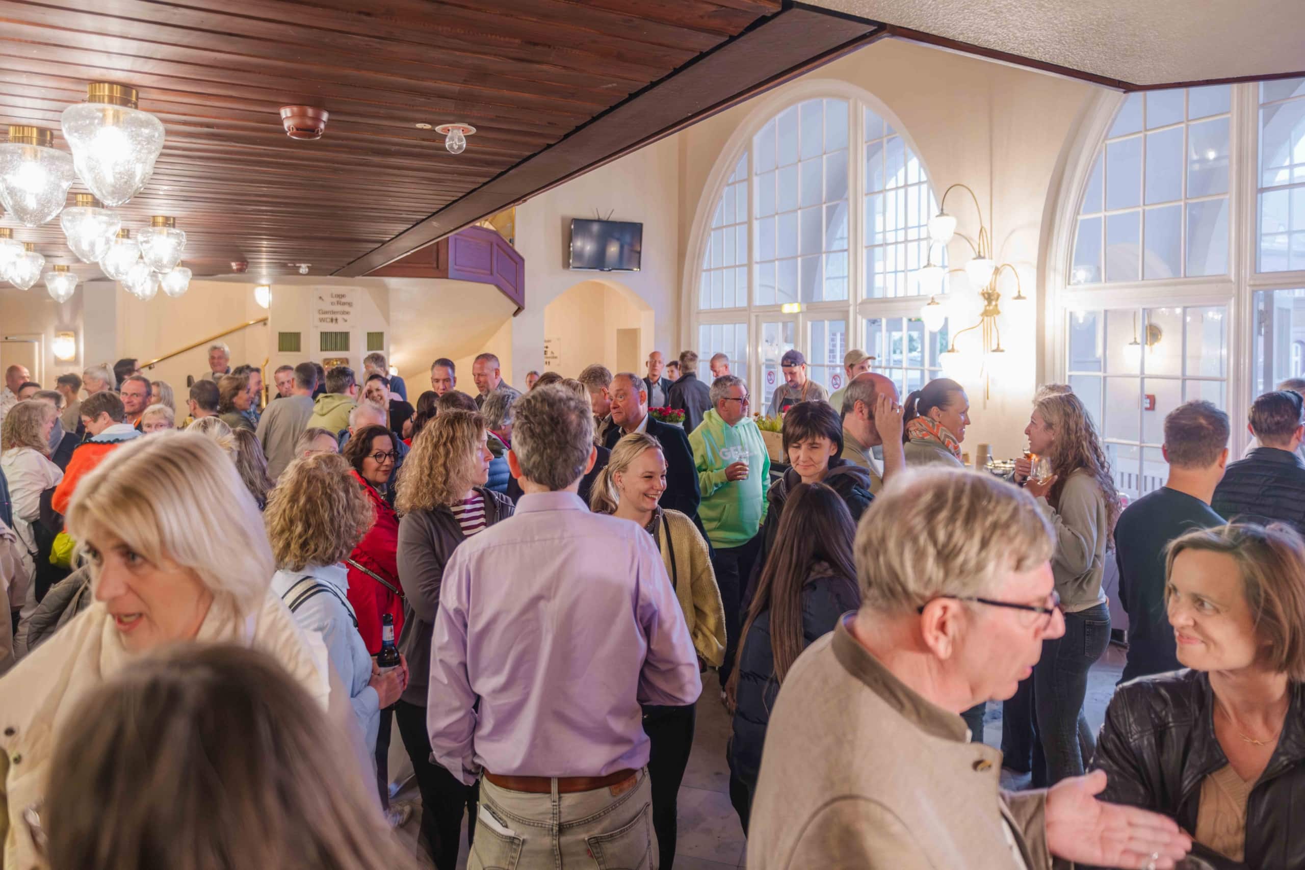 Eine große Gruppe von Menschen unterhält sich in einem hellen, geräumigen Raum mit Bogenfenstern und Kronleuchtern. Viele unterhalten sich in kleinen Gruppen, was eine lebhafte, freundliche Atmosphäre schafft.