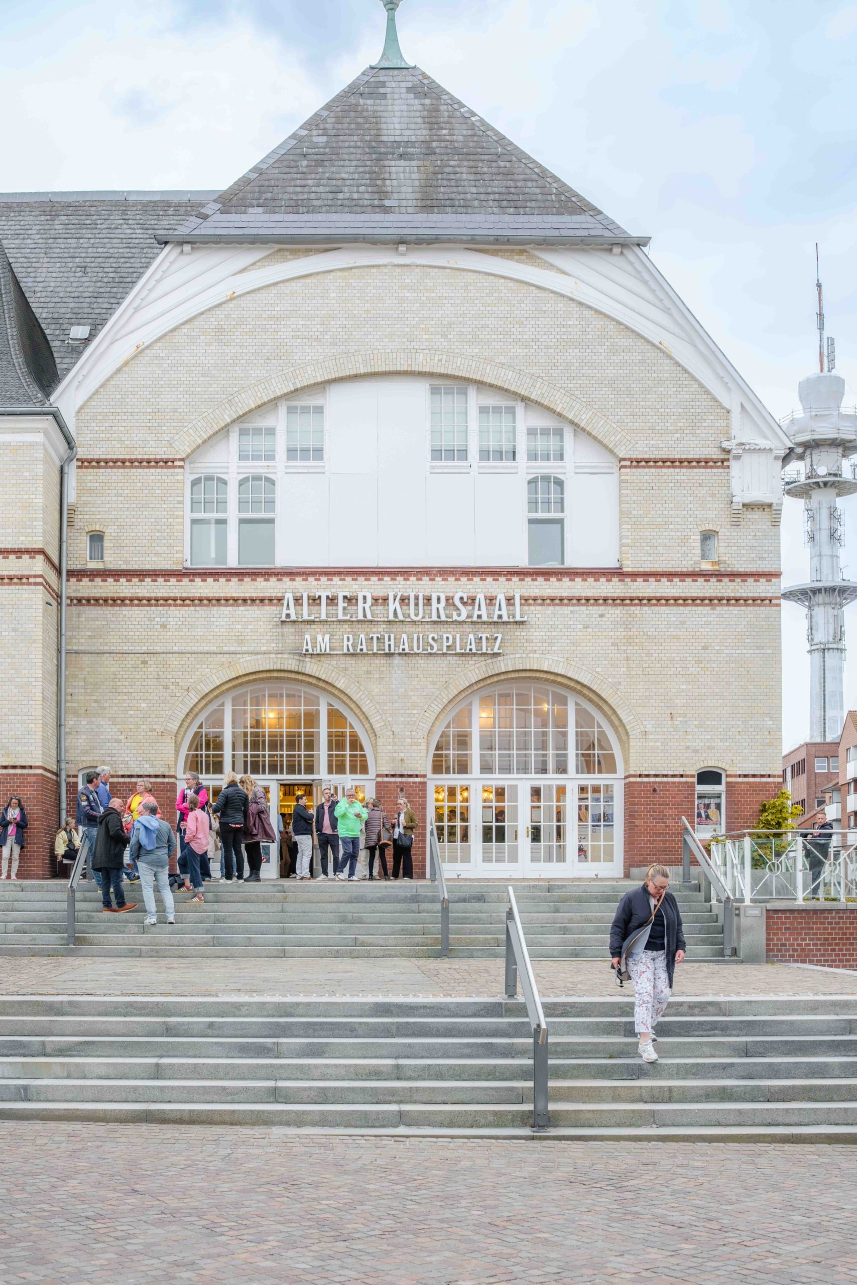 Eine Gruppe von Menschen steht vor dem historischen Gebäude „Alter Kursaal am Rathausplatz“, das große Bogenfenster und eine Backsteinfassade aufweist. Im Vordergrund geht eine Person die Treppe hinunter.
