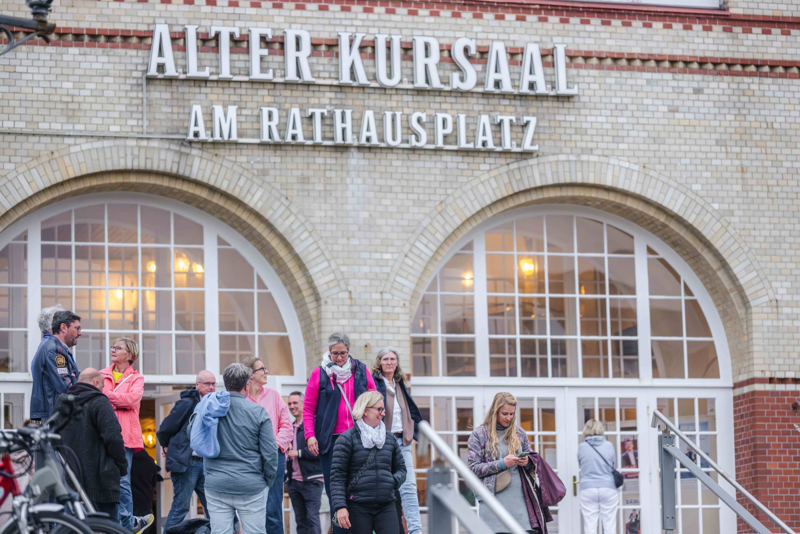 Vor dem Eingang eines Gebäudes, über dessen großen Bogenfenstern und -türen ein Schild mit der Aufschrift „Alter Kursaal am Rathausplatz“ angebracht ist, steht eine Gruppe von Menschen und geht hin und her.