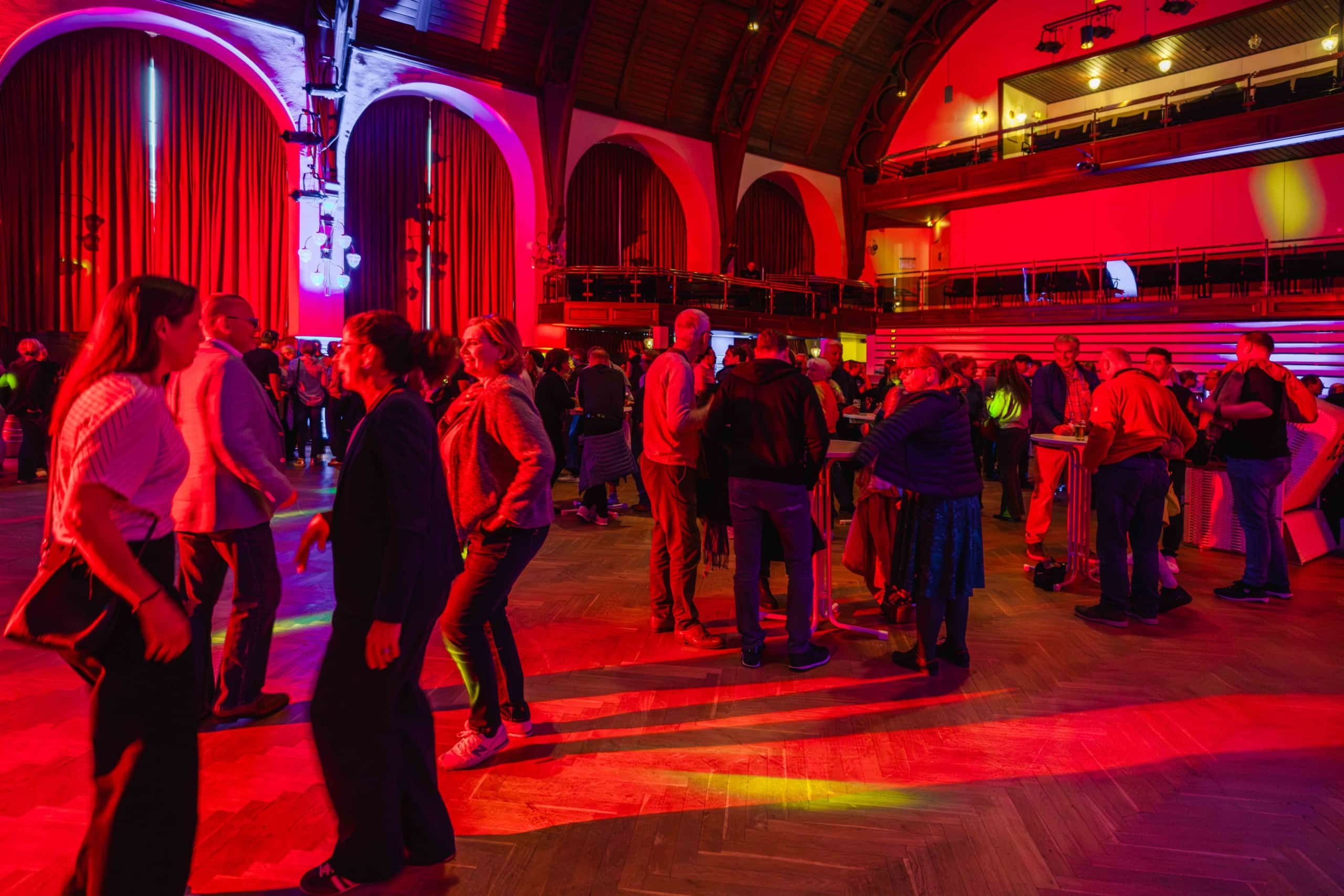 In einem großen Saal mit hohen Decken, roter Beleuchtung und Bogenfenstern versammeln sich die Menschen und unterhalten sich. Einige tanzen, während andere in Gruppen stehen oder an Tischen sitzen. Die Atmosphäre wirkt lebhaft und festlich.